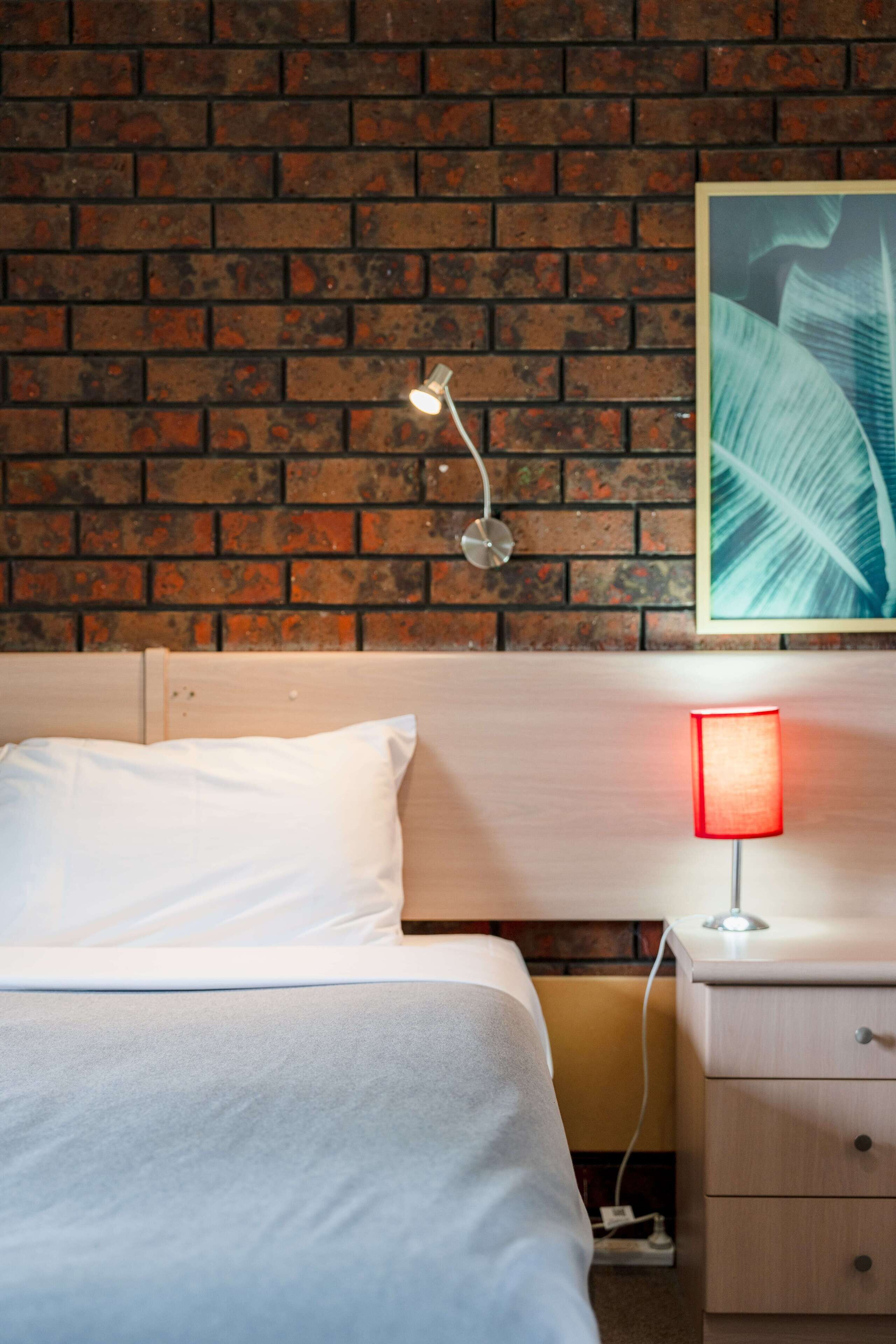 Una habitación de hotel con una pared de ladrillo, una cama con ropa blanca, una mesita de noche con una lámpara roja y un cuadro en la pared.