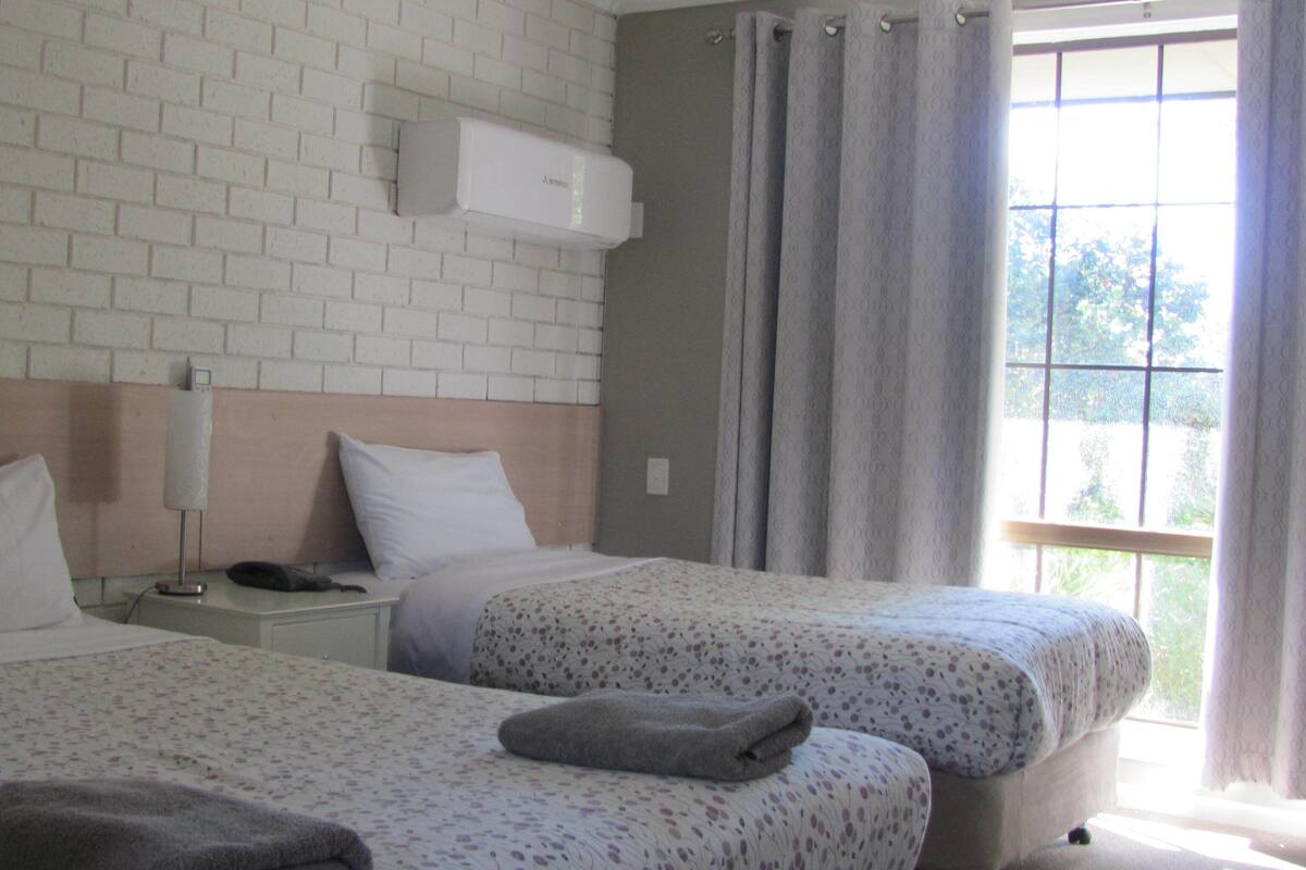 A hotel room with two beds, a brick wall, an air conditioning unit, a nightstand, a lamp, and a window with curtains.