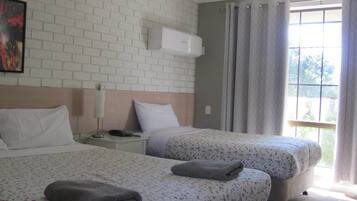 A hotel room with two beds, a brick wall, an air conditioning unit, a nightstand, a lamp, and a window with curtains.