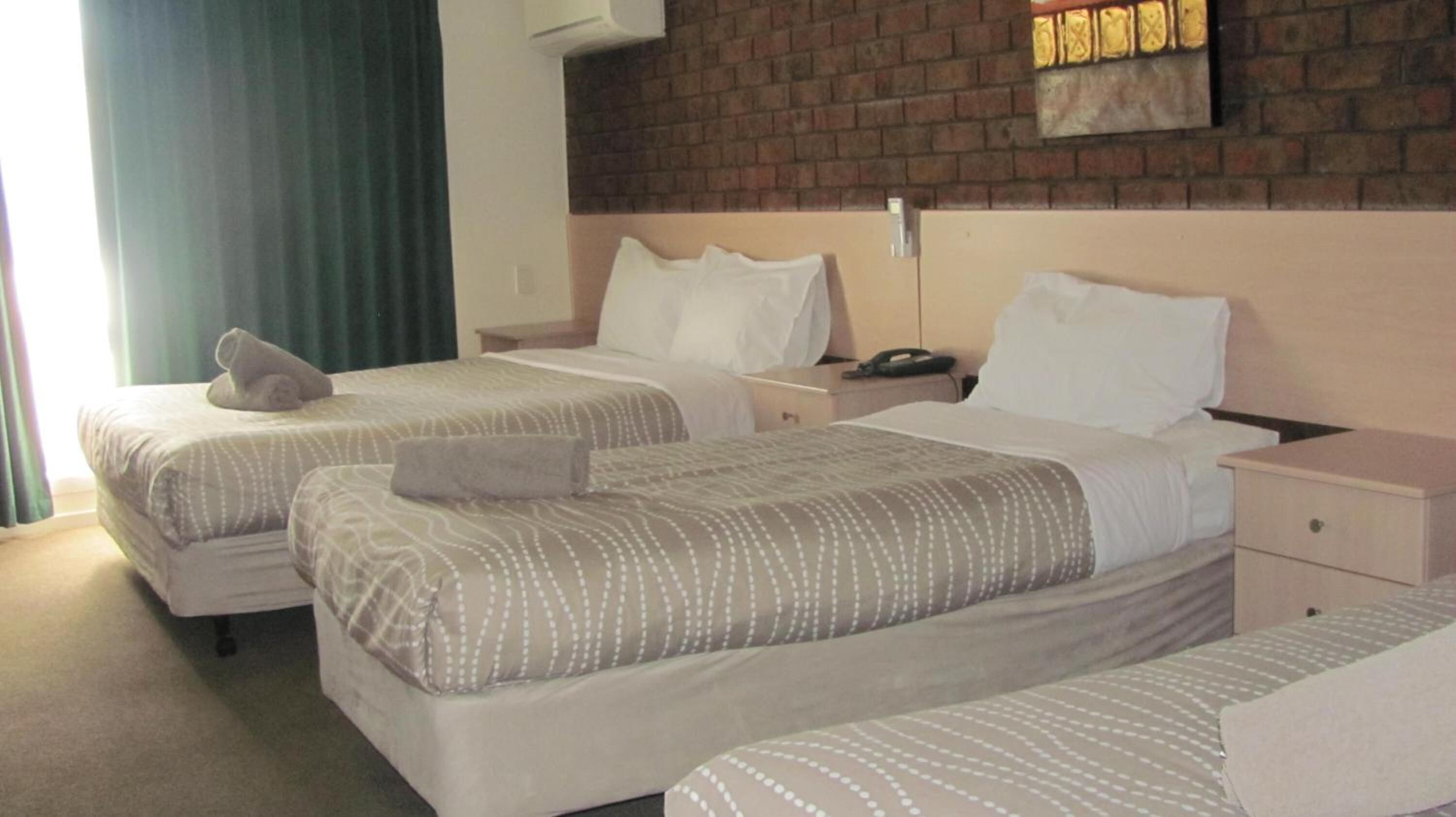 A hotel room with two beds, each with a pillow and a blanket. There is a nightstand with a phone and a lamp. The room has a brick wall and a window with curtains.