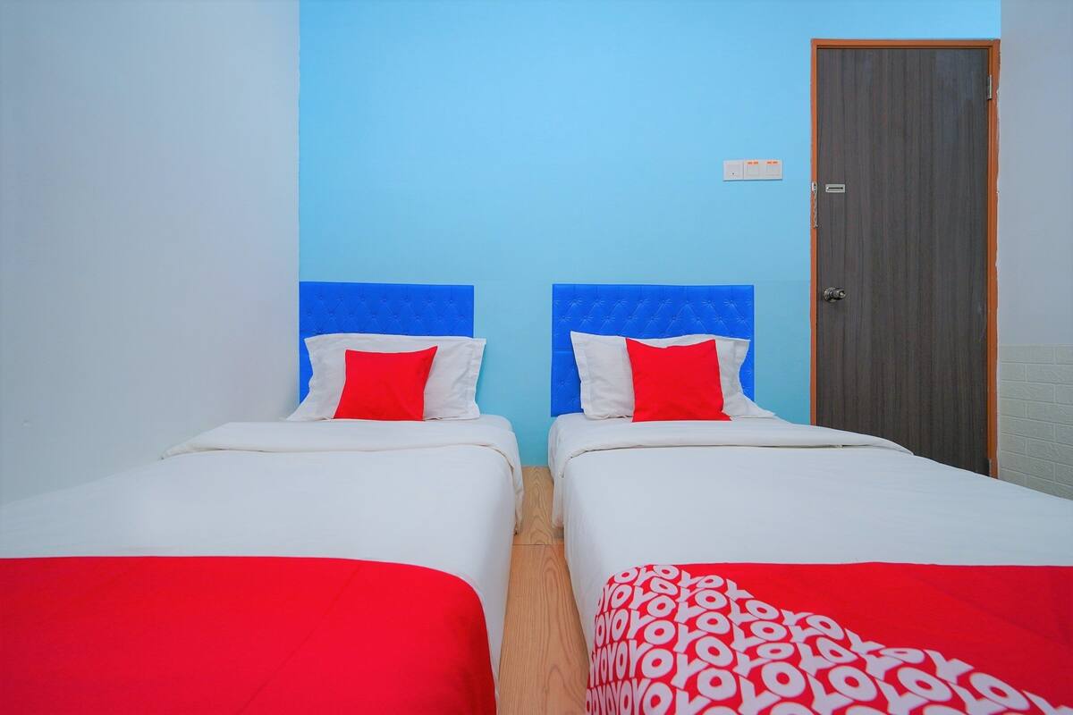 Une chambre avec deux lits, chacun doté d’une tête de lit bleue, de draps blancs et d’oreillers rouges. On y trouve une porte en bois et une petite pancarte accrochée au mur.
