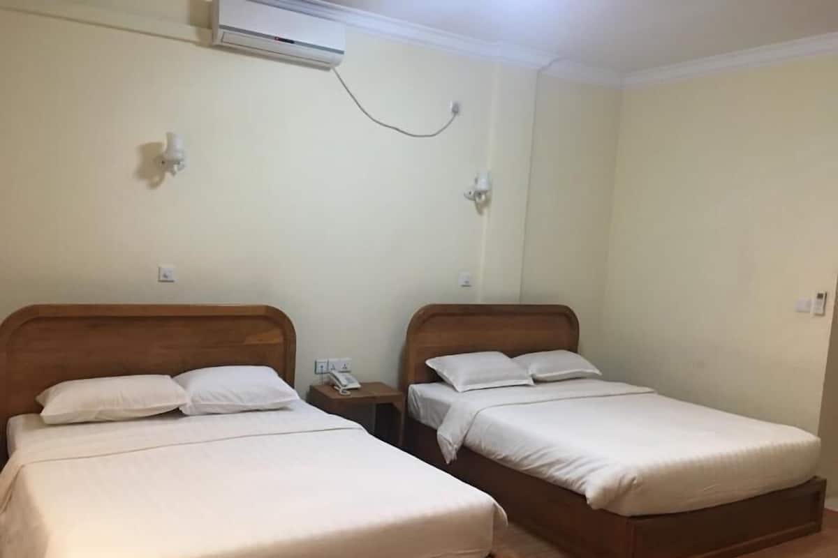 A room with two single beds, each with a wooden headboard and white bedding. There is a wall-mounted air conditioner above the beds and a small bedside table with a phone.
