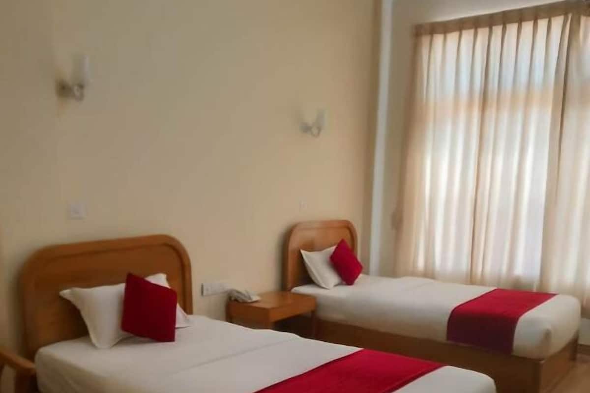 Two single beds with wooden headboards, white bedding, and red accents. There are wooden nightstands and a small table. The room has beige walls and a large window with curtains.