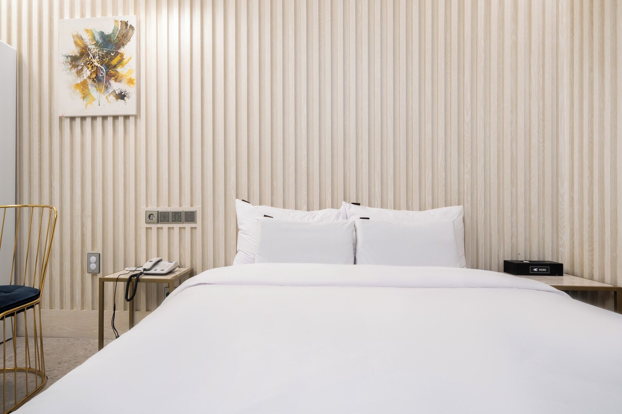 A hotel room with a bed, a chair, a bedside table with a phone, and a wall-mounted artwork.