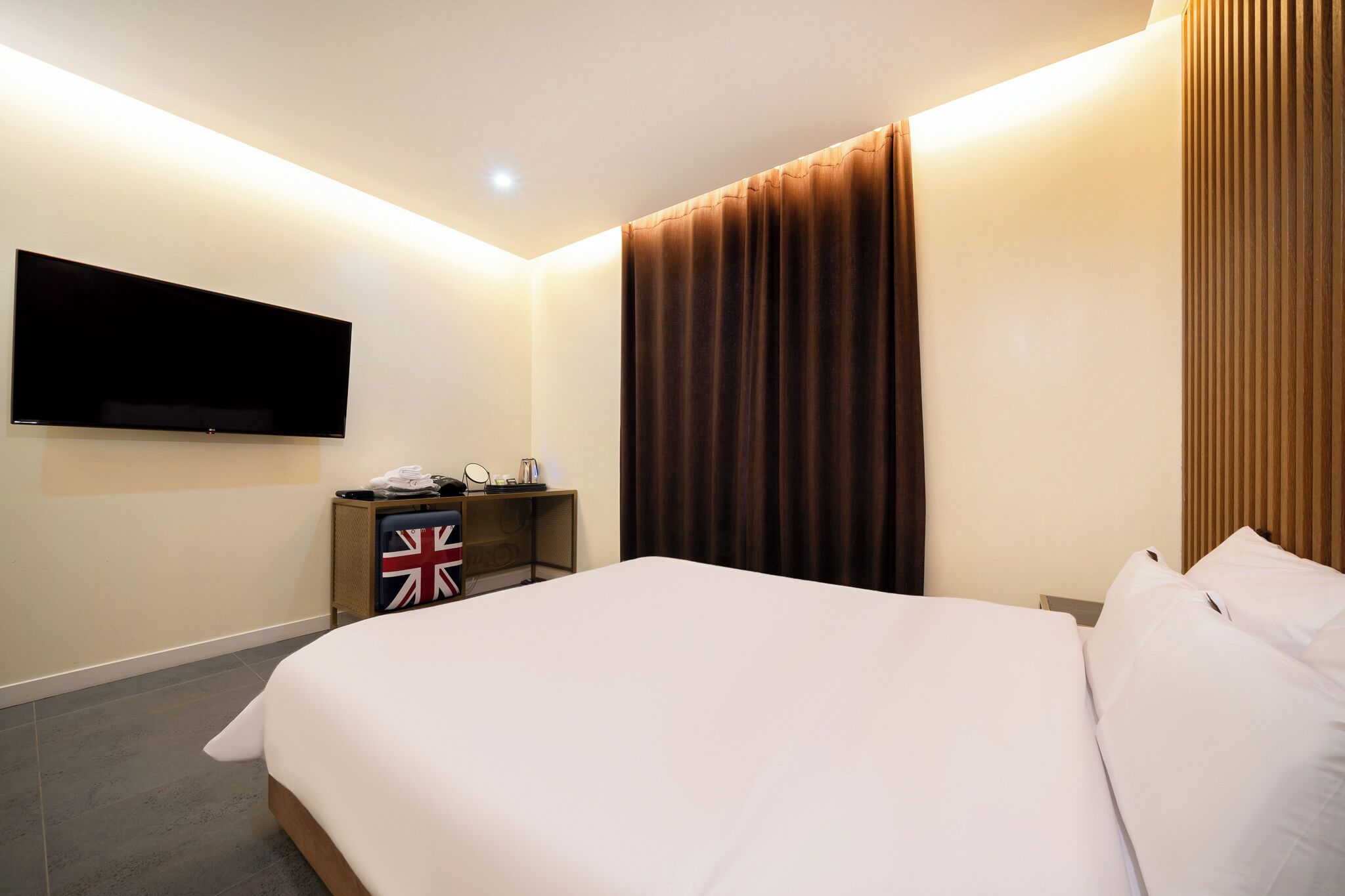 Habitación de hotel con una cama grande, un televisor de pantalla plana montado en la pared, una mesita de noche con un teléfono y una bandera, y una cortina en la ventana.