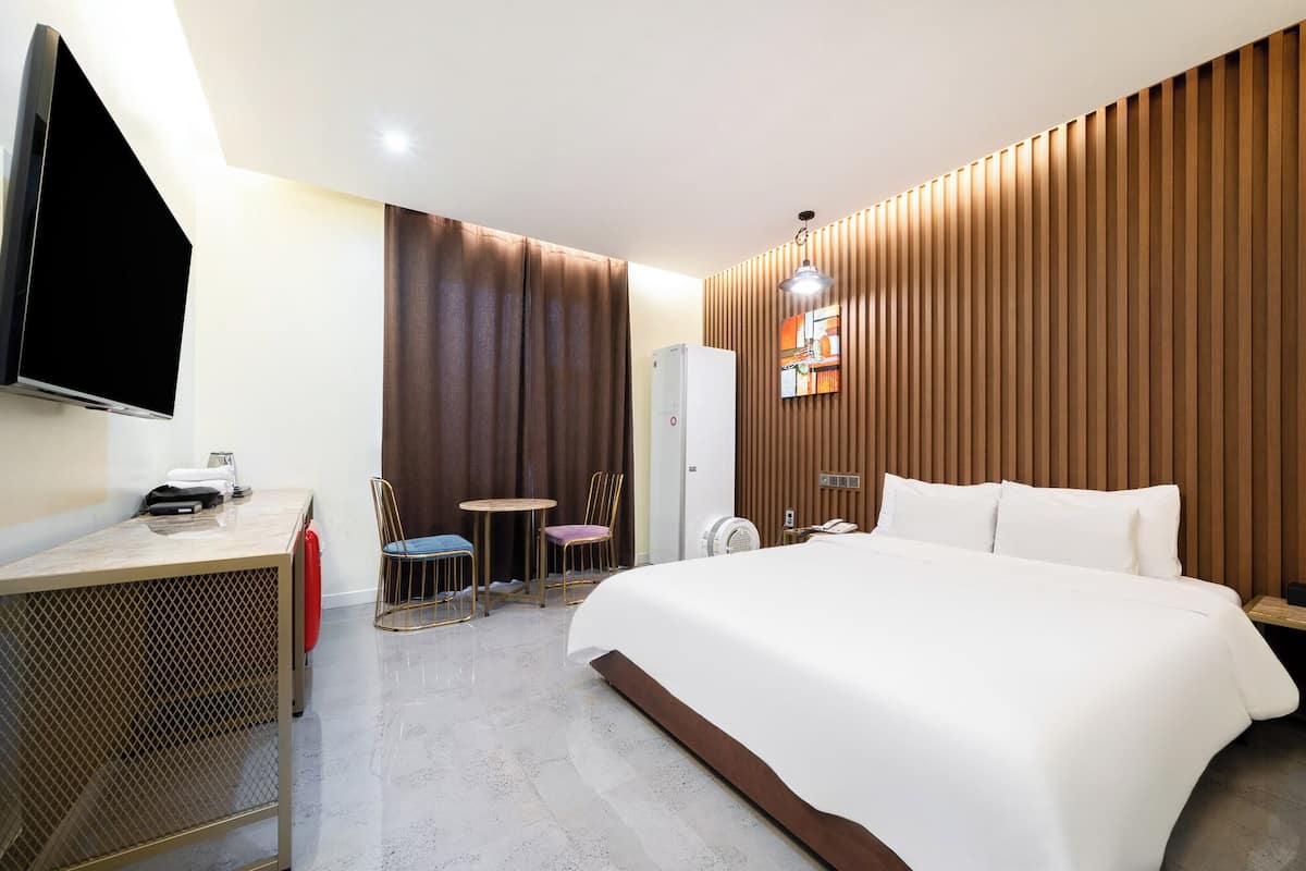 A hotel room with a large bed, a desk with a flat-screen TV, a small round table, two chairs, and a wardrobe.