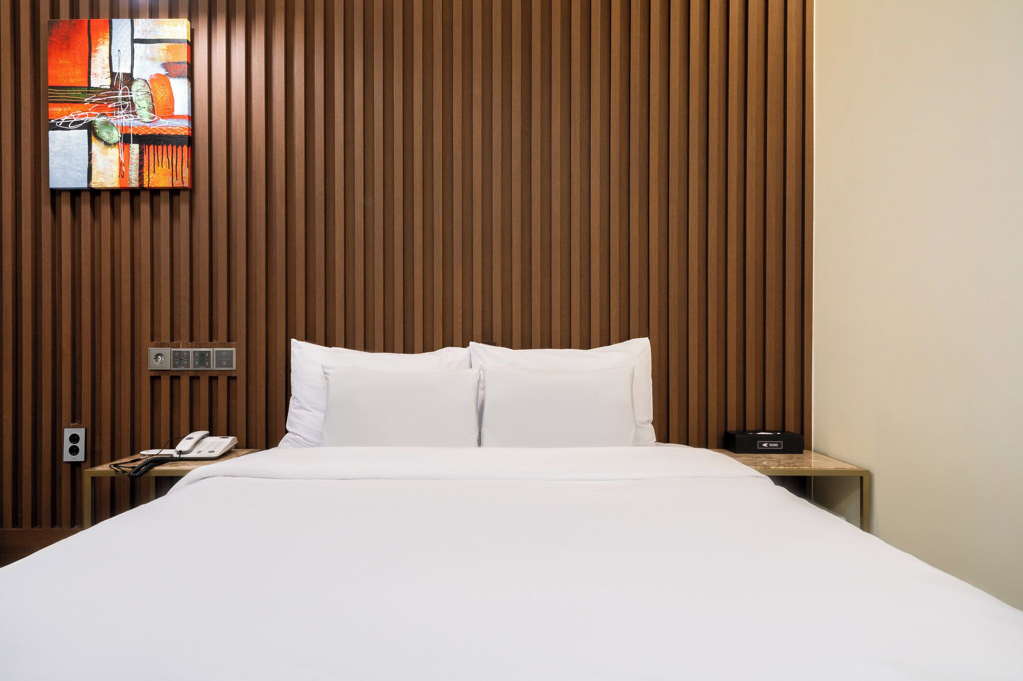 Habitación de hotel con pared de listones de madera, cama con ropa blanca, mesita de noche con teléfono y radio, y un cuadro sobre la cama.