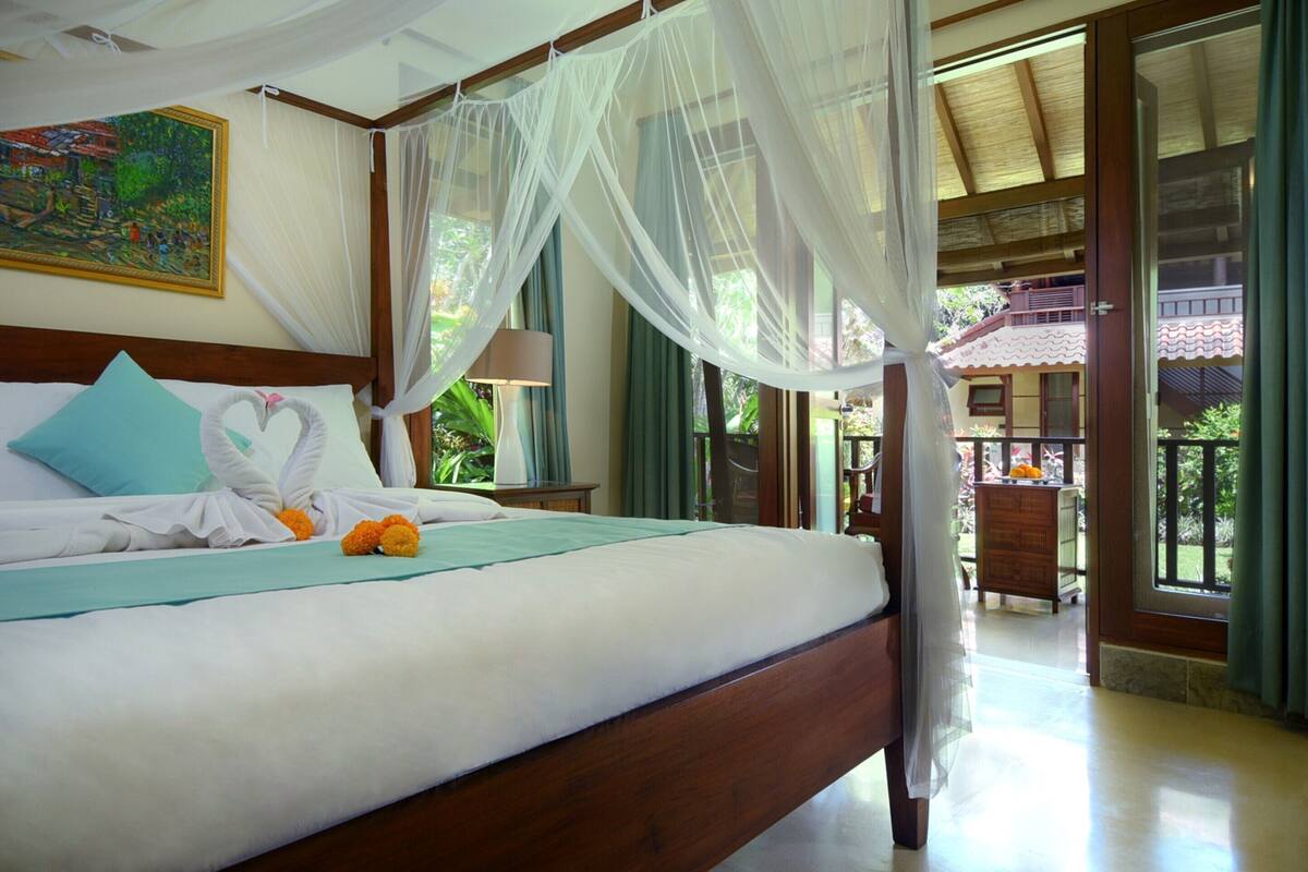 A bedroom with a bed, a nightstand, a lamp, a painting, and a view of the outside.