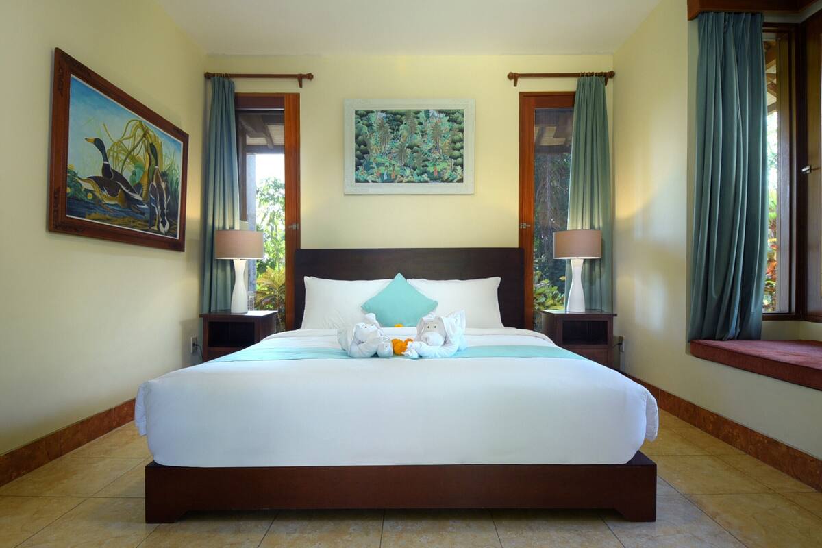 A large bed with a white bedspread, two nightstands with lamps, a painting on the wall, and a window with curtains.