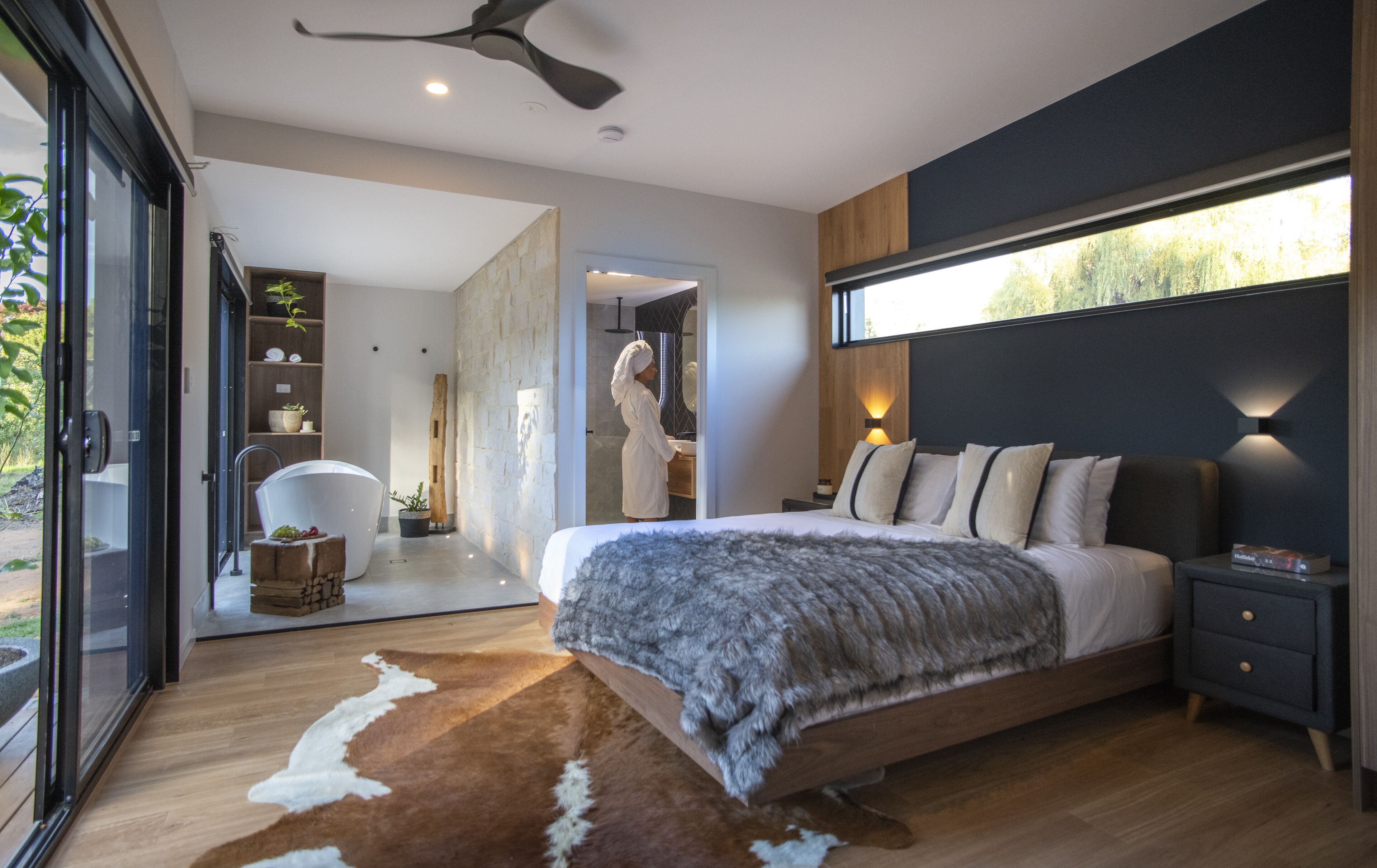 A modern bedroom with a large bed, a wooden floor, a cowhide rug, a nightstand, and a large window with a view of the outdoors.