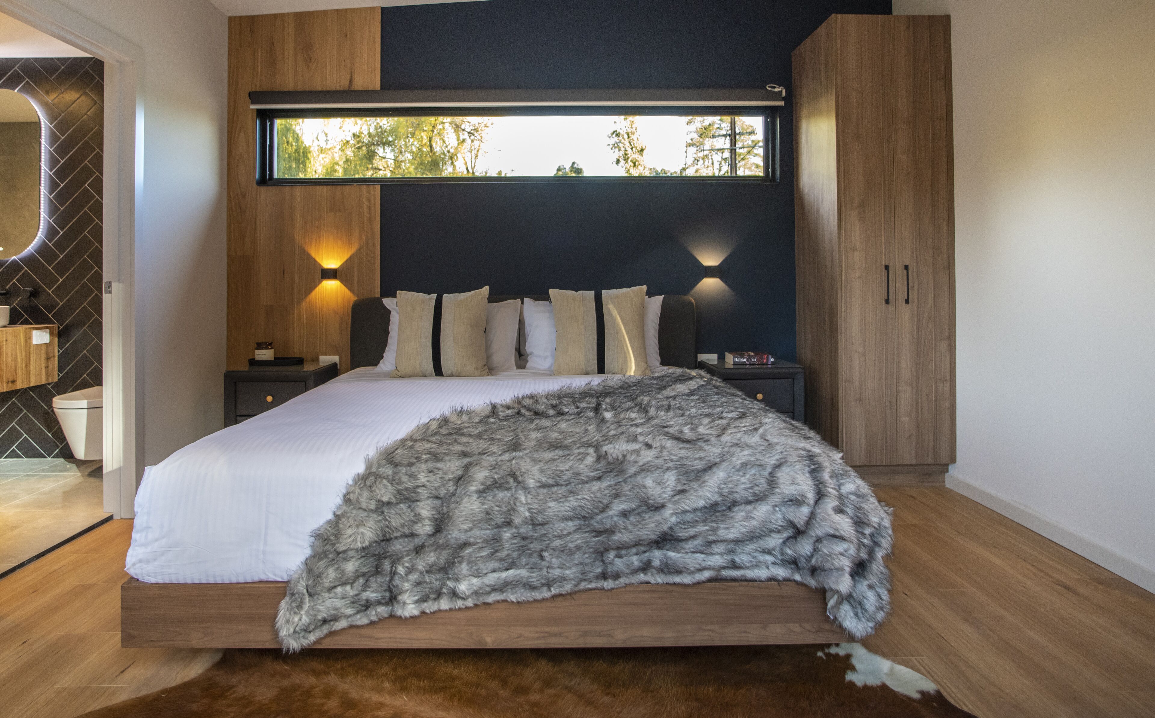 A modern bedroom with a large bed, wooden headboard, and a view of trees through a window.
