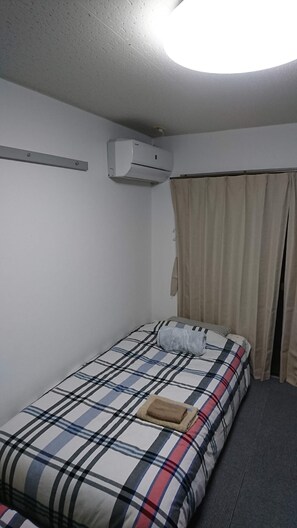 A small, single-bed room with a plaid bedspread, an air conditioning unit mounted on the wall, and a curtain on the window. - Uzumasa 203 (Kyoto)