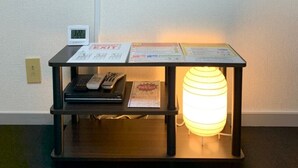 Una TV a schermo piatto montata sulla parete sopra un tavolino consolle in legno con una lampada accesa, una sveglia digitale e alcuni documenti.