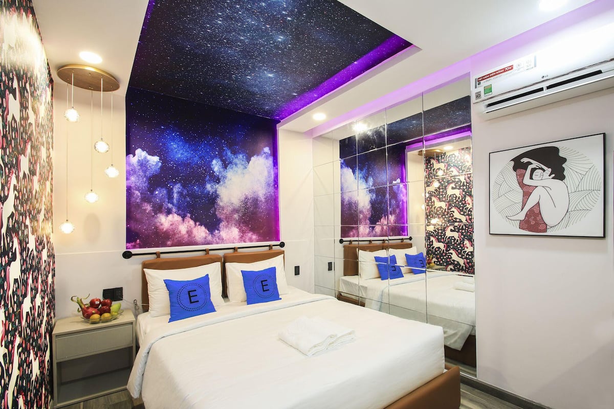 A modern bedroom with a starry sky mural, a bed with blue pillows, a fruit bowl on the nightstand, and a large mirror reflecting the room.