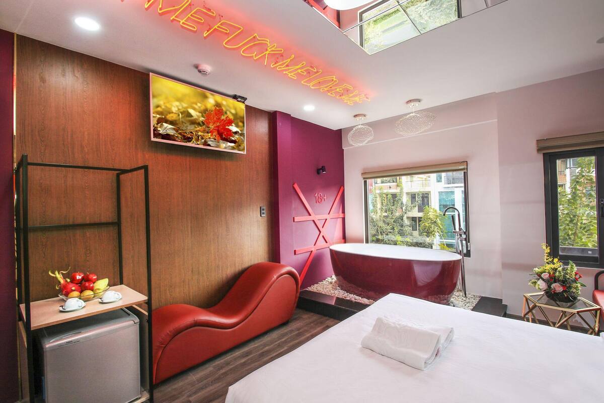 A modern hotel room with a large bed, a red massage chair, a small fridge, a red bathtub, and a large window offering a view of the outside.