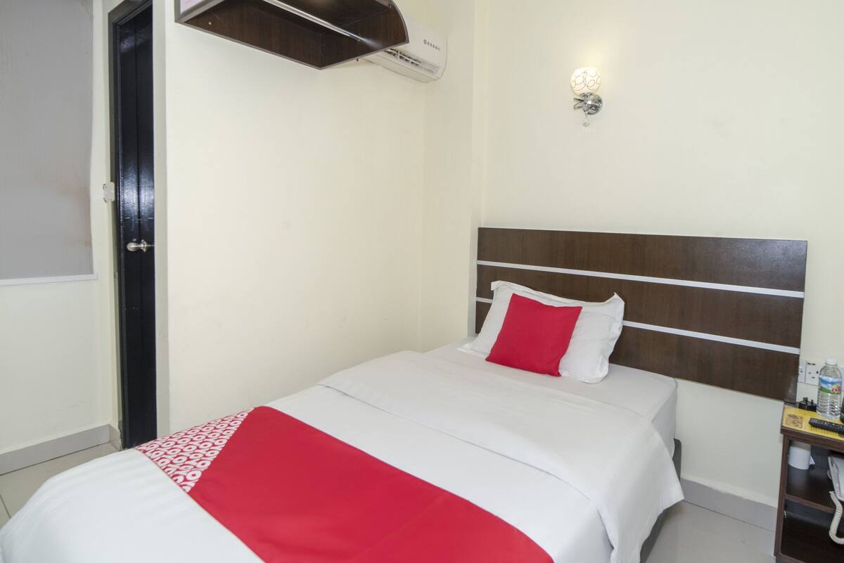 A hotel room with a bed, a bedside table with a bottle and a remote control, a wall-mounted air conditioner, and a door with a window.