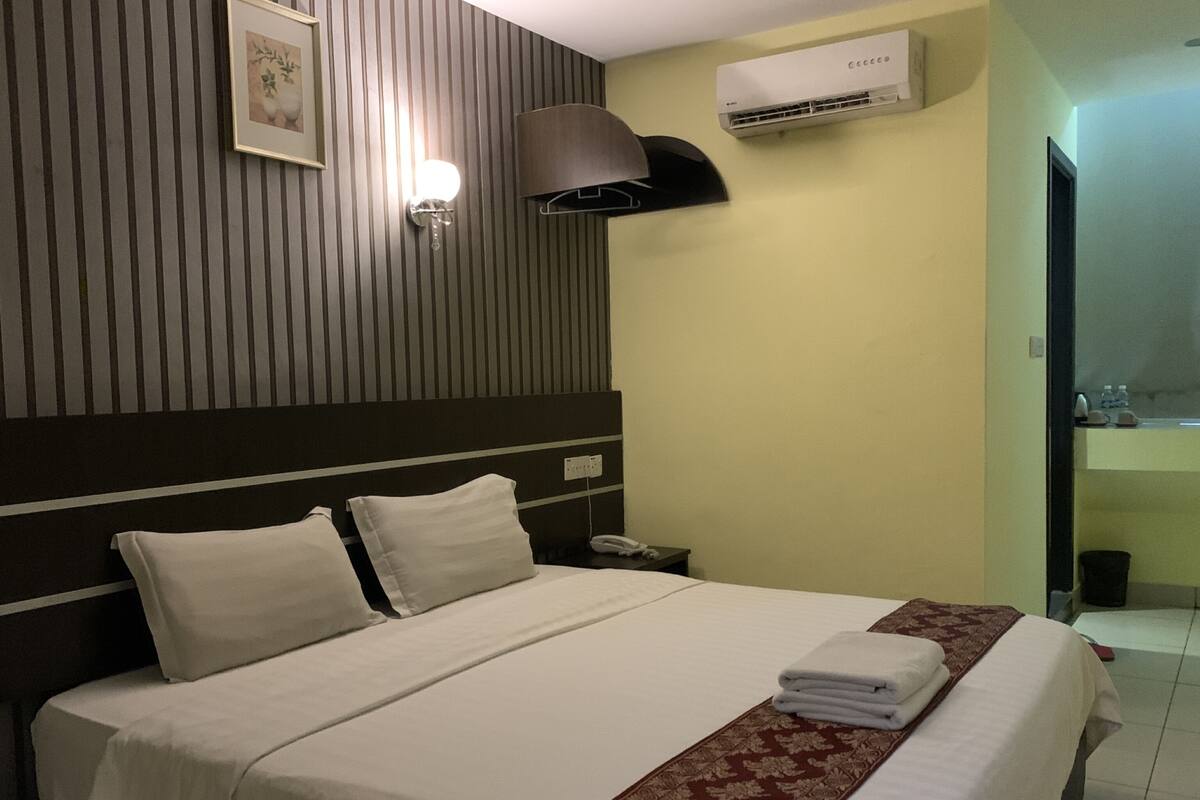 A hotel room with a bed, a nightstand, an air conditioning unit, and a wall-mounted lamp.