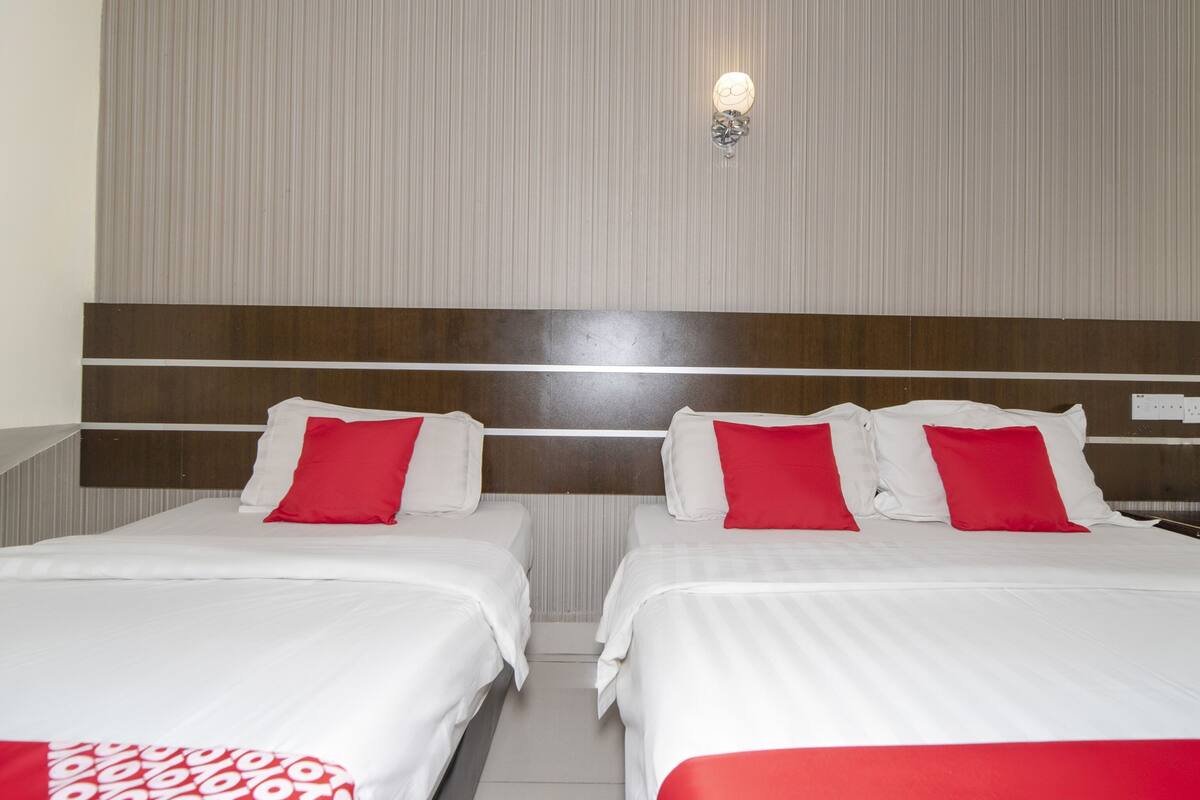 A hotel room with two beds, each with white bedding and red pillows. There is a wall-mounted light fixture above the beds.