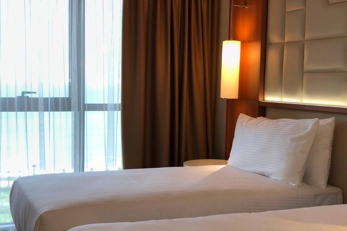 A hotel room with two beds, a large window, and a wall-mounted lamp.