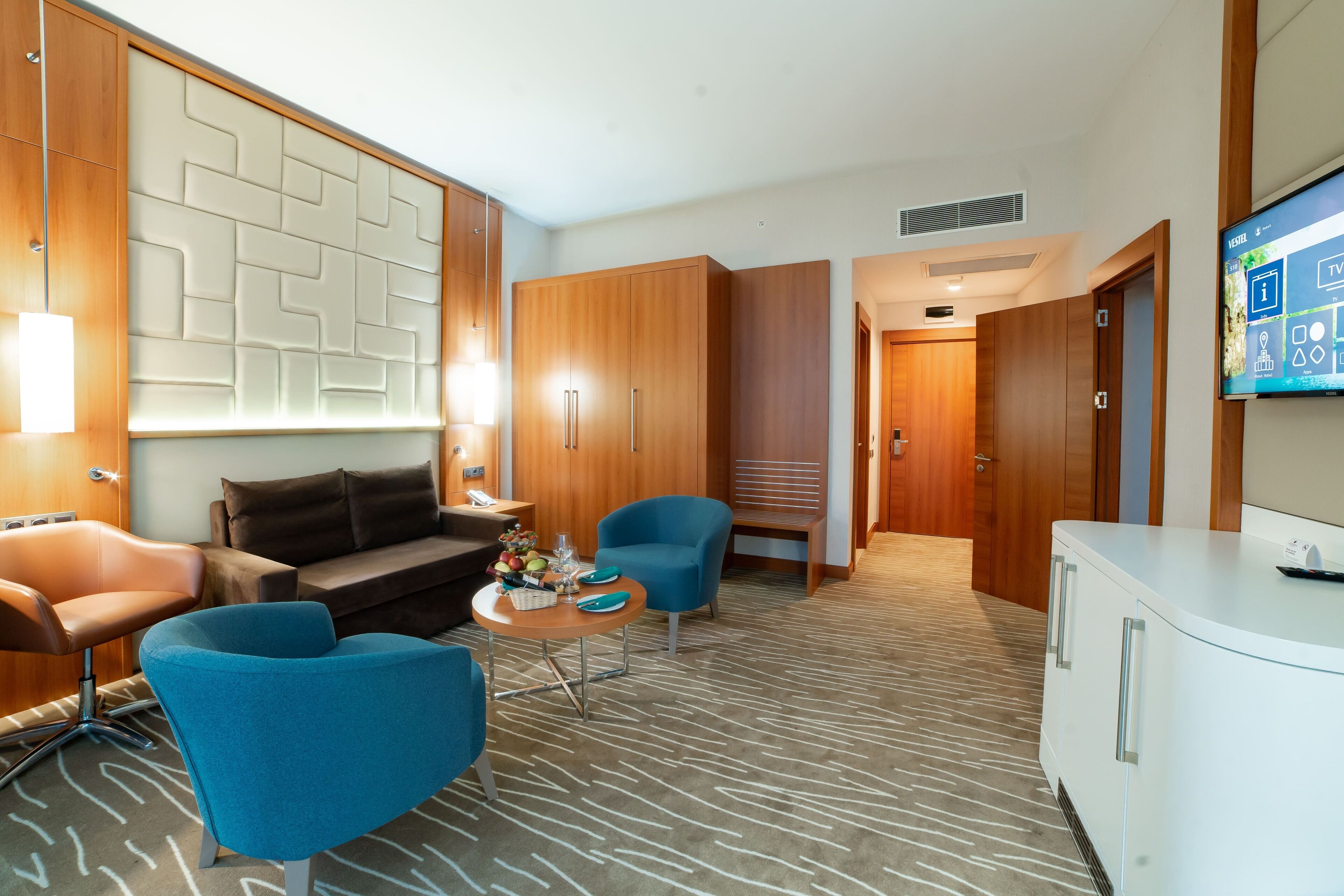 A hotel room with a modern design, featuring a sofa, two armchairs, a small round table with a fruit bowl, and a TV mounted on the wall.