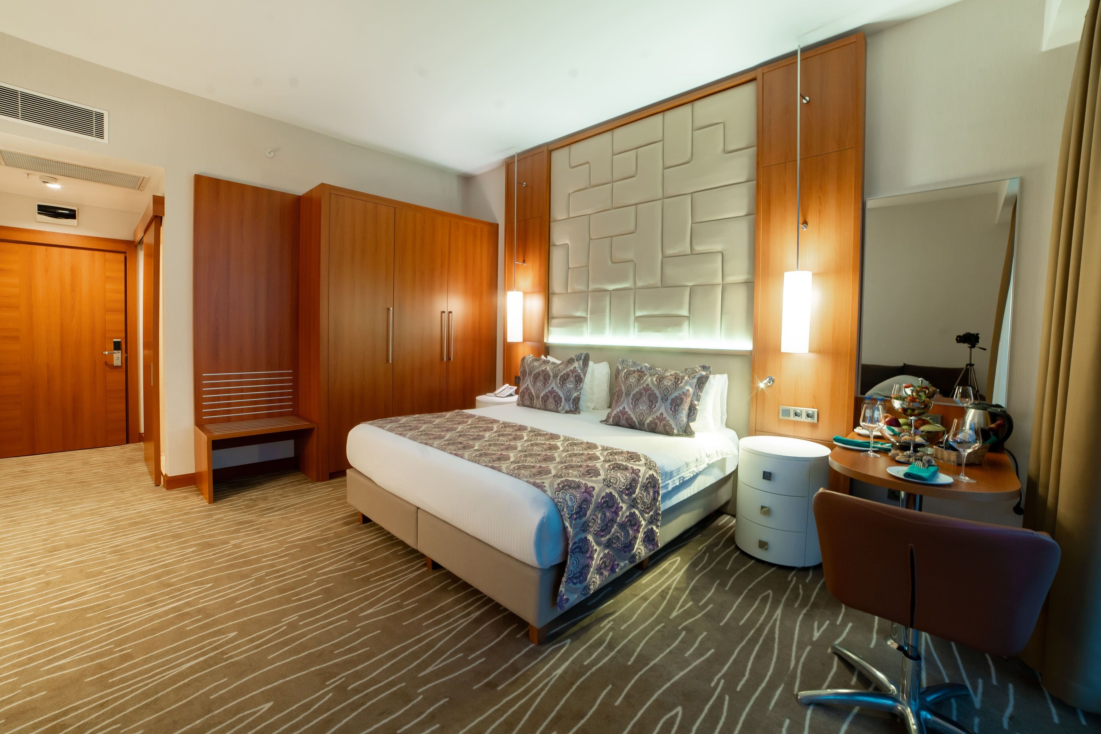 A hotel room with a bed, bedside table, mirror, wardrobe, and a desk with a chair.