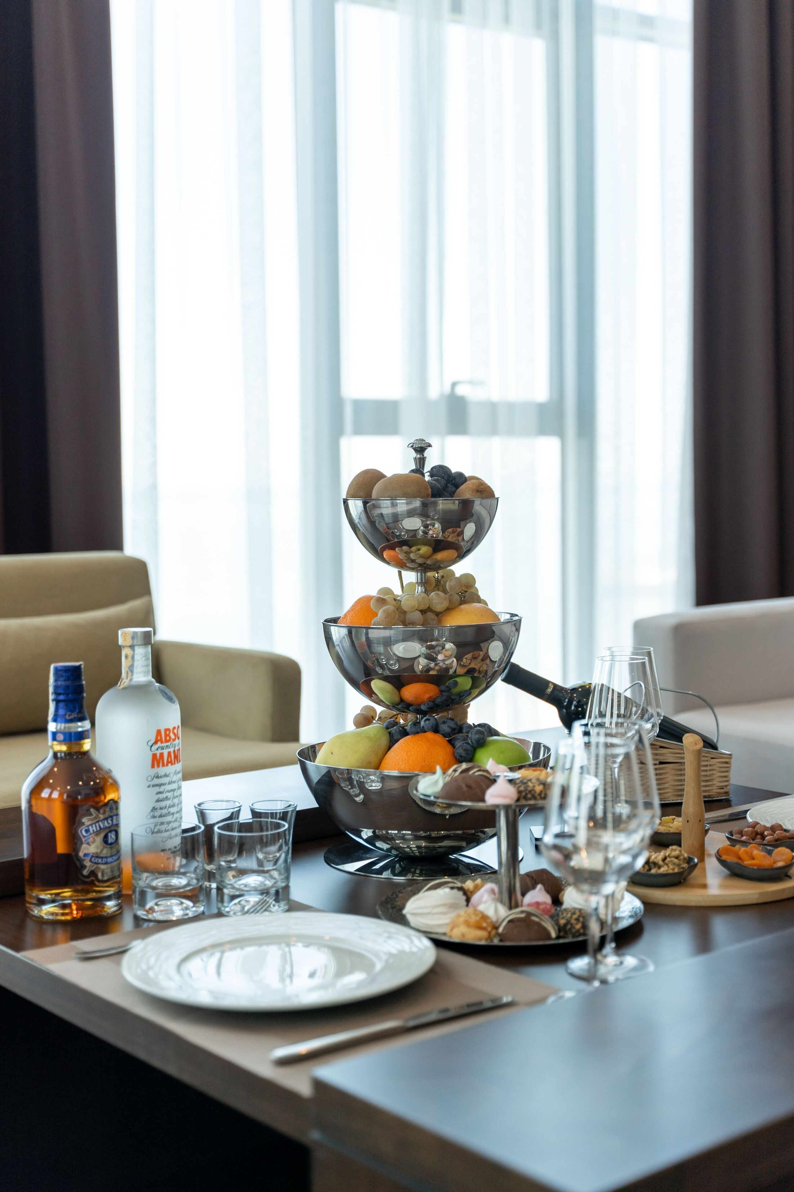 Une table à manger avec un présentoir à fruits et à fruits secs à trois niveaux, une bouteille d’Absolut et des verres à vin.