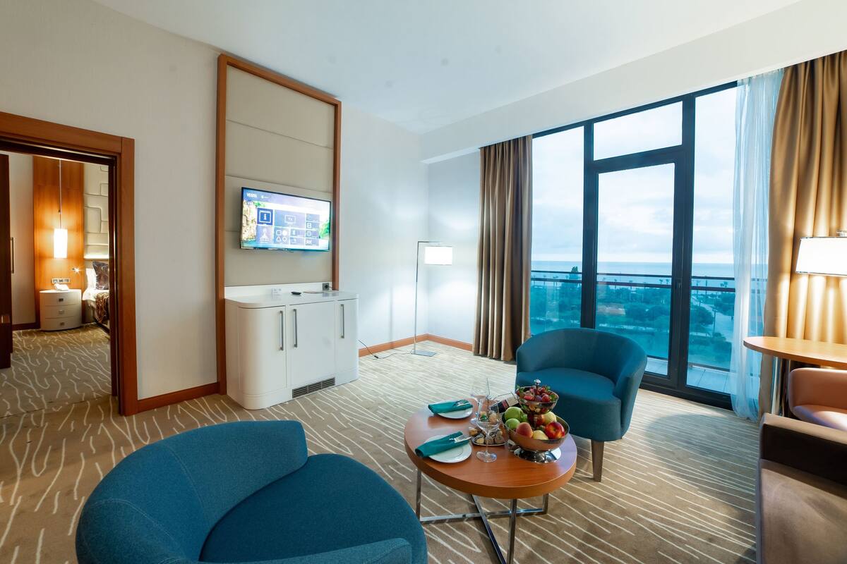 A hotel room with a large window, a flat-screen TV, a dining table with fruit, two blue armchairs, and a minibar.