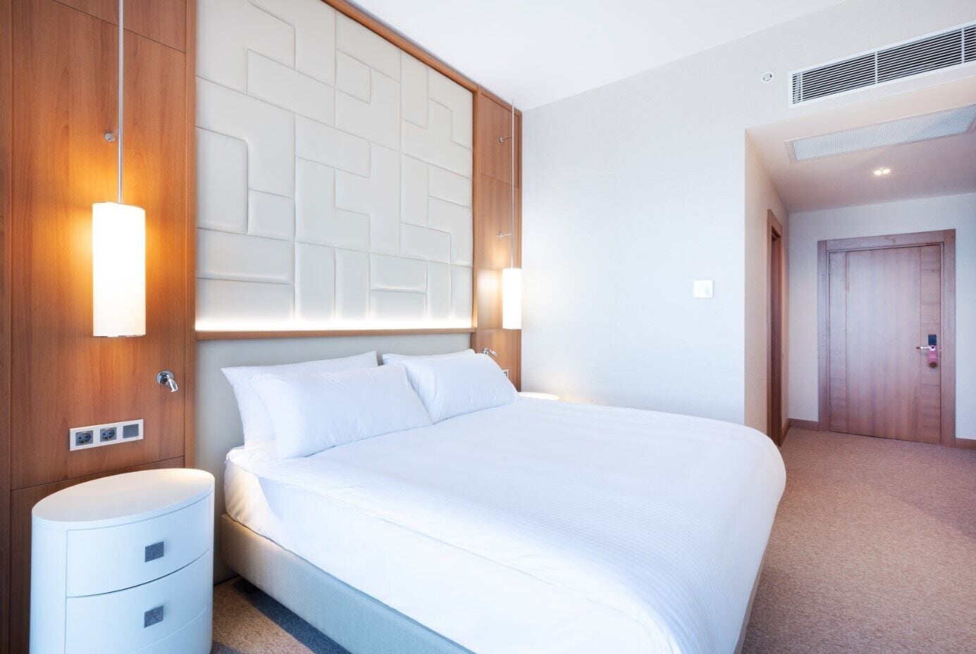 Ein modernes Hotelzimmer mit einem großen Bett, einem Holzpaneel-Kopfstück, Nachttischlampen und einem kleinen Nachttisch.