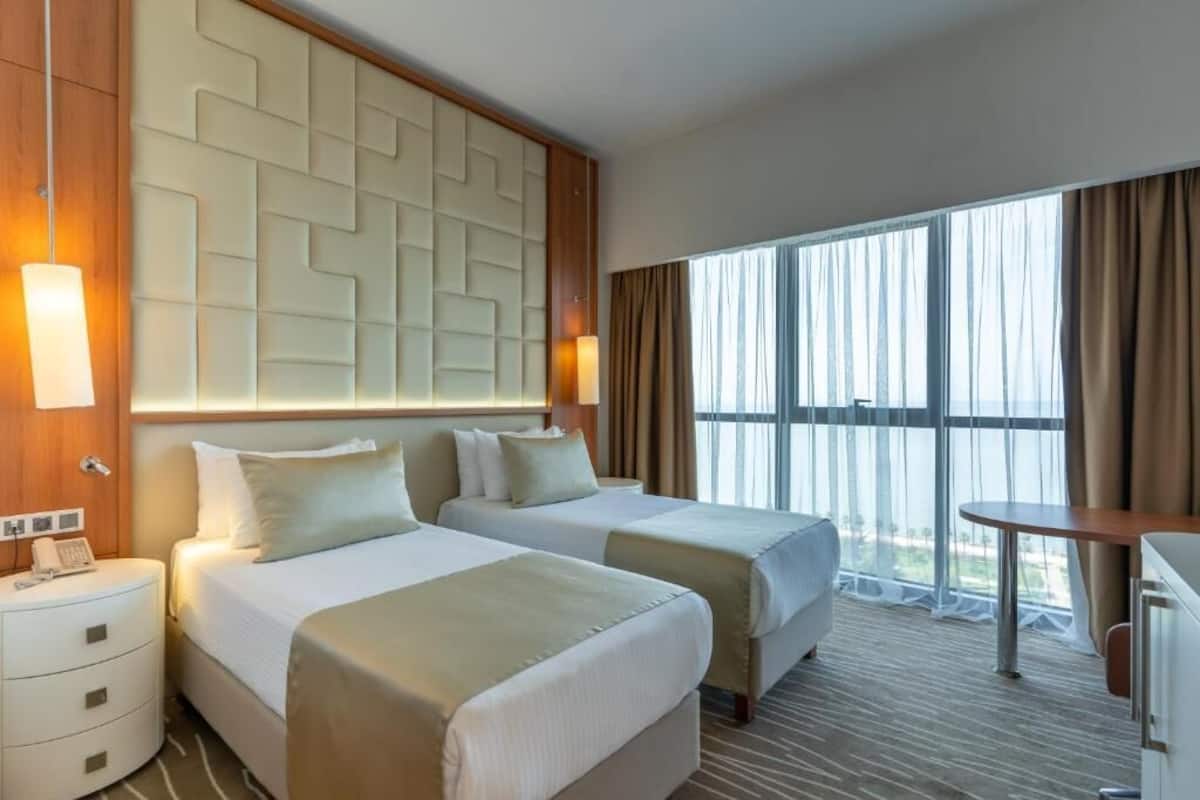 A hotel room with two beds, a desk, a chair, a large window with curtains, and a wall with a geometric pattern.