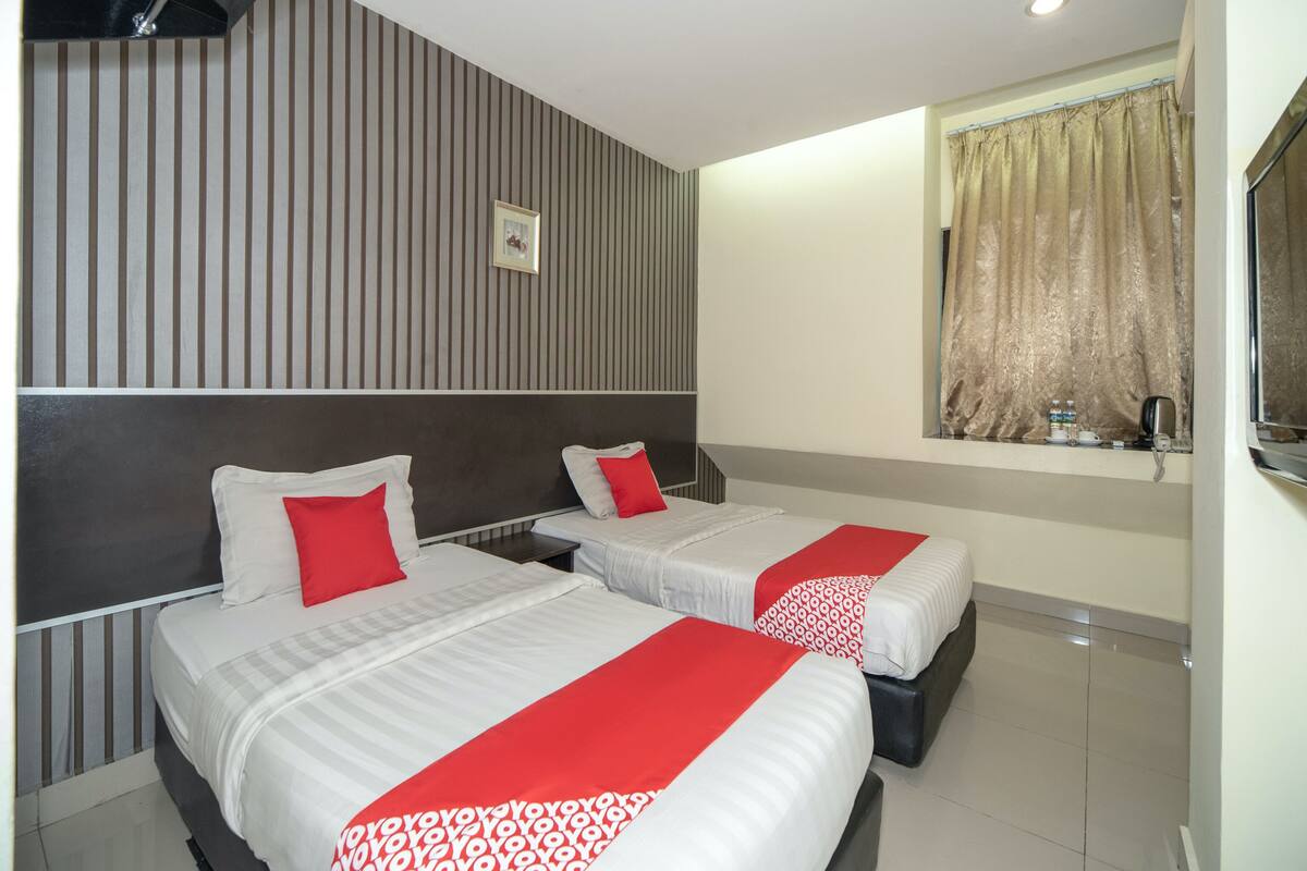 A hotel room with two beds, striped wallpaper, a window with curtains, and a small table with a kettle and cups.