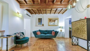 A room with a herringbone wooden floor, a teal sofa with pink cushions, a round table with a lamp, and a large floral artwork on the wall. - Skylife Spanish Steps Apartment (Roma)