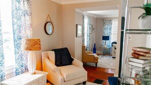 En stue med en hvid sofa, et sofabord, en lampe og et mønstret gardin. Der er en stol og et lille bord med en lampe i baggrunden.