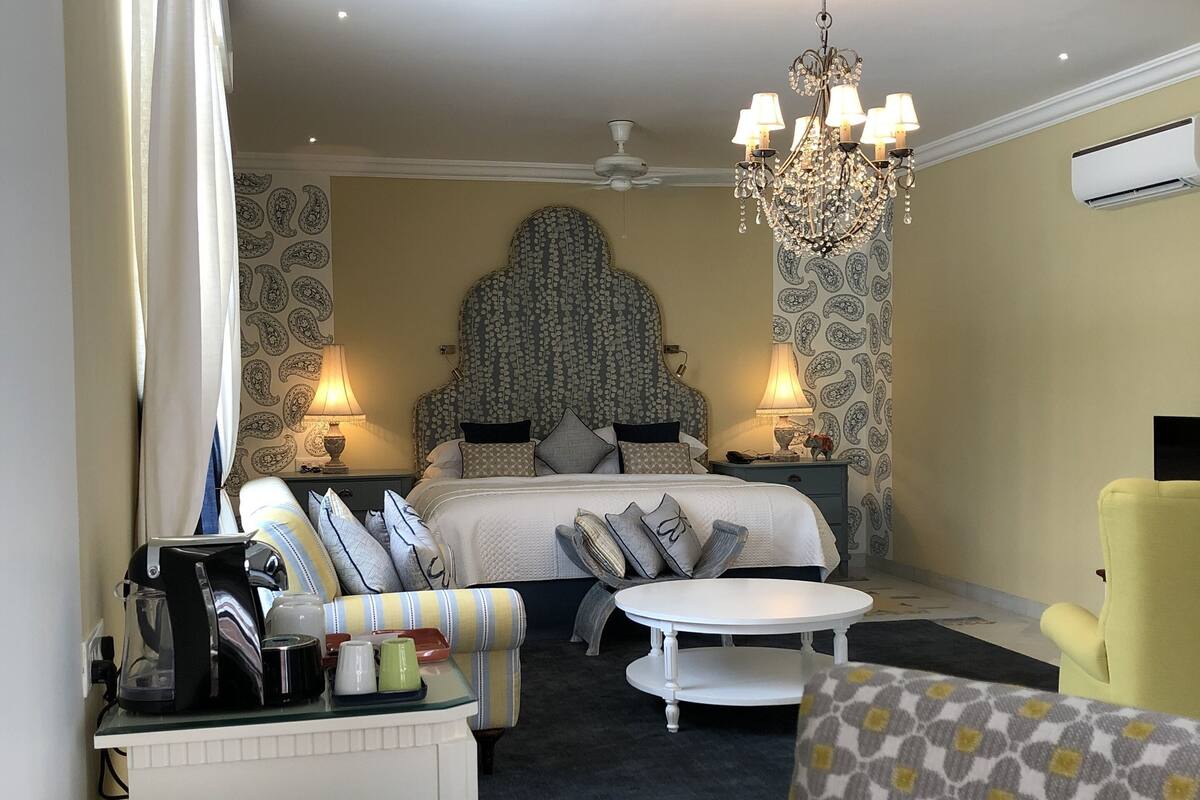 A hotel room with a bed, a chandelier, a desk with a lamp, and a chair.