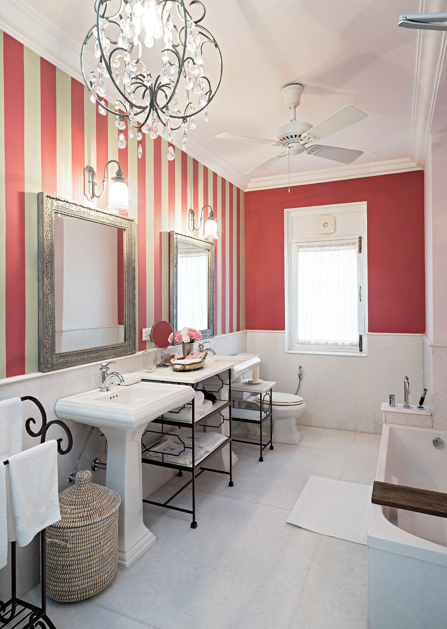 Une salle de bain aux murs rayés, ornée d’un lustre, d’un ventilateur de plafond, d’un grand miroir, d’une baignoire et d’un meuble-lavabo avec un évier et un porte-serviettes.