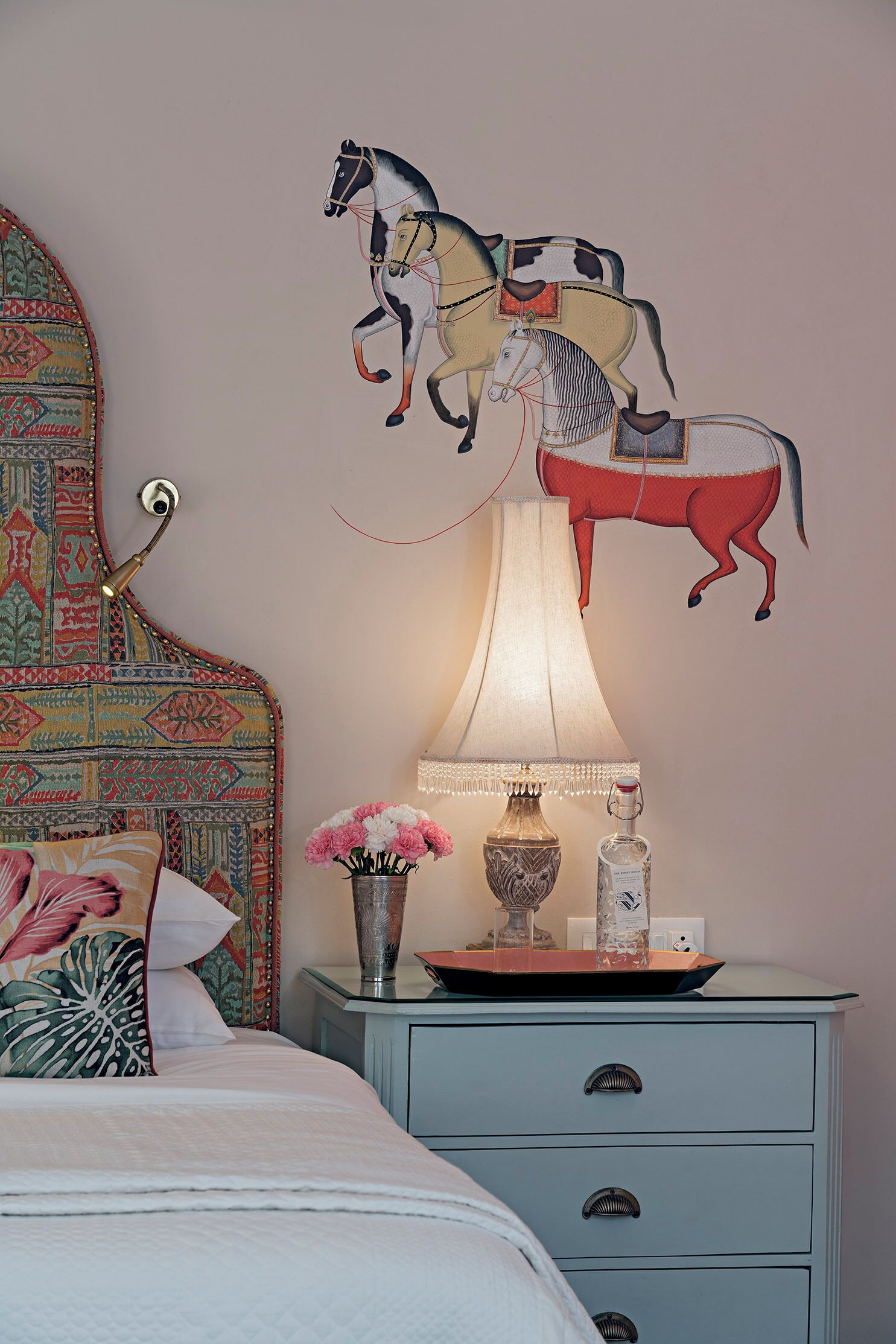 Un dormitorio con cama, mesita de noche, lámpara, un jarrón con flores y un cuadro de un caballo en la pared.