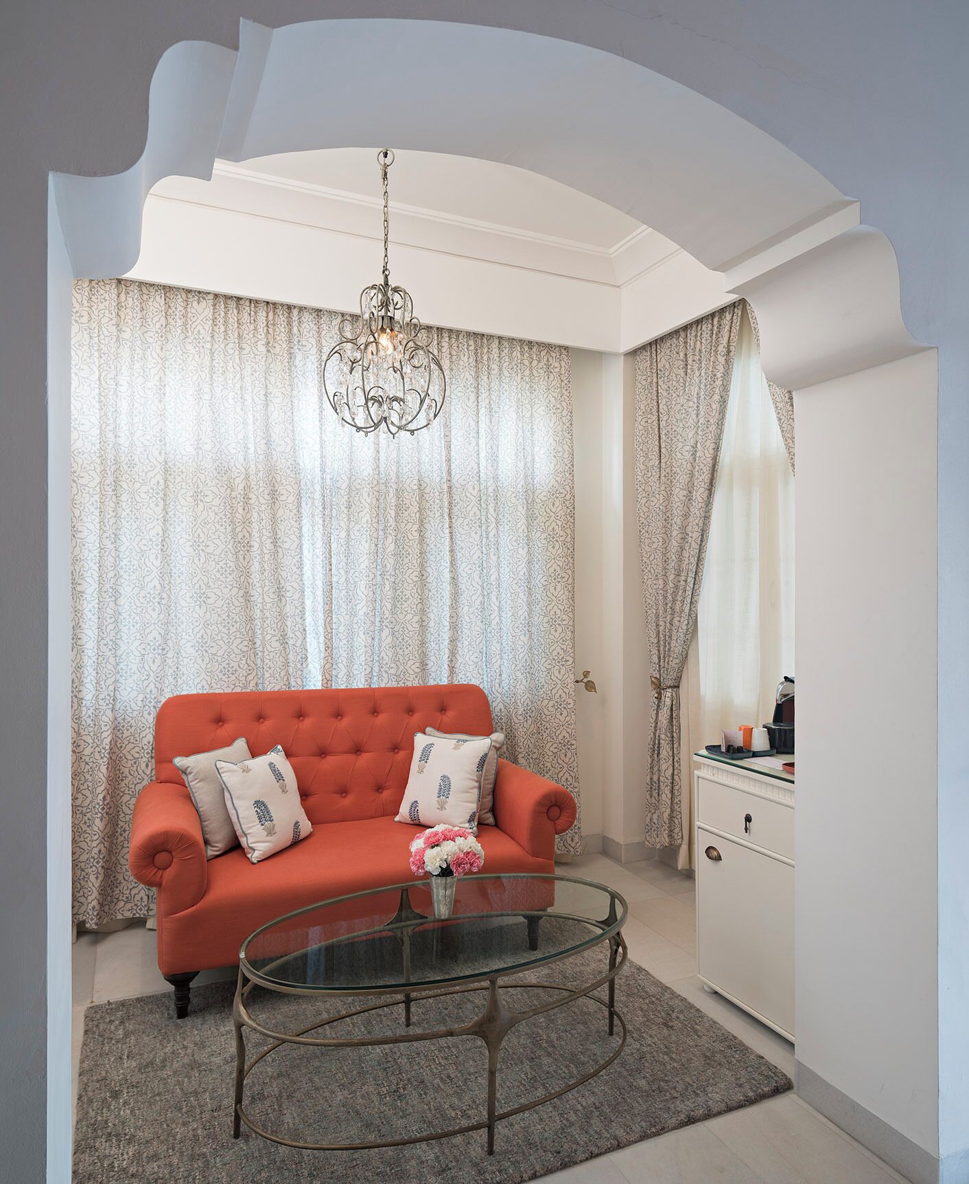 Una sala de estar moderna con un sofá rojo, cortinas estampadas, una mesa de centro de vidrio y una araña.