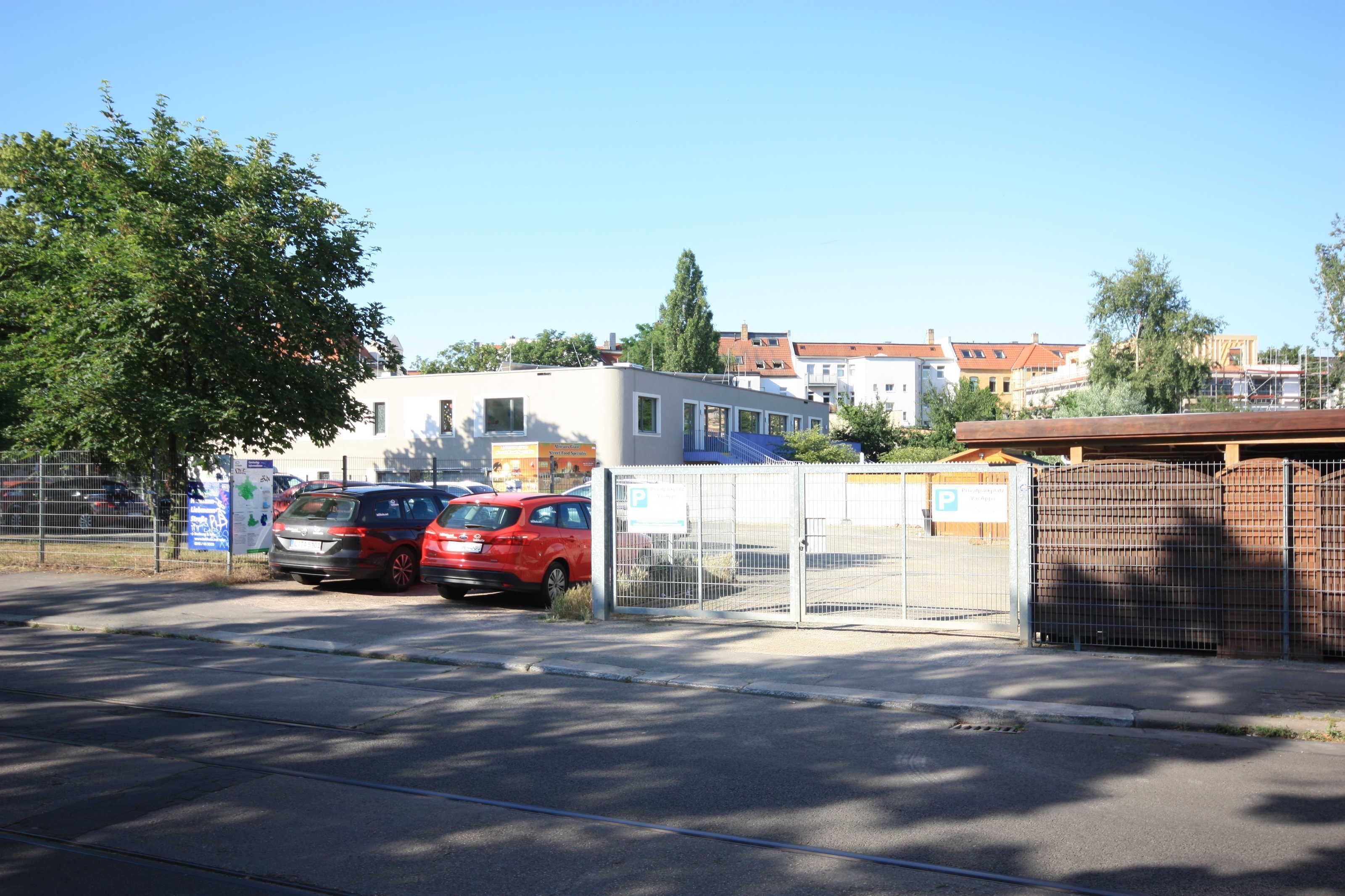 Ein Parkplatz mit zwei Autos, einem Zaun und Gebäuden im Hintergrund.