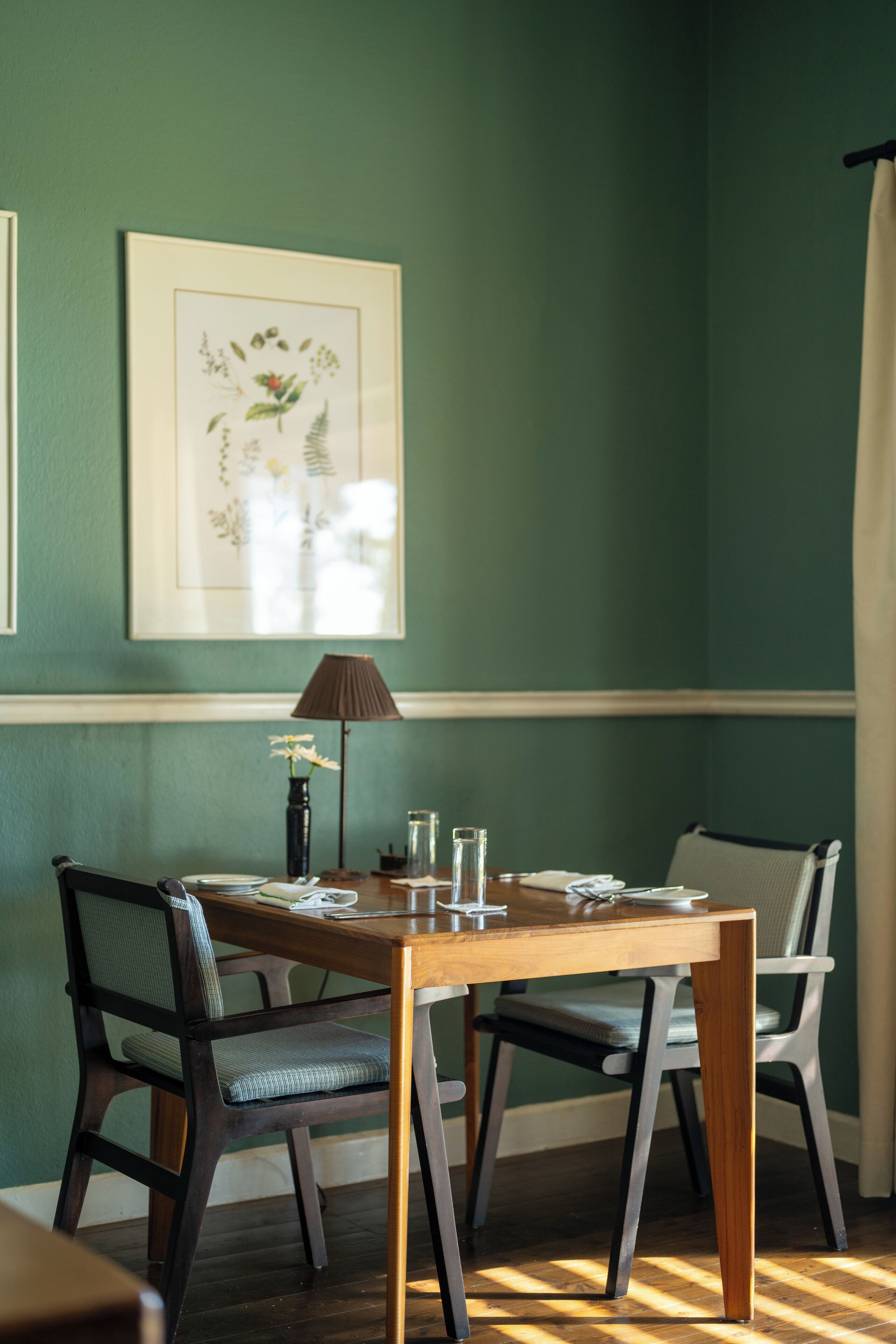 Ein Essbereich mit Holztisch, zwei Stühlen, einer Blumenvase, einer Lampe und gerahmten Pflanzenmotiven an der Wand.