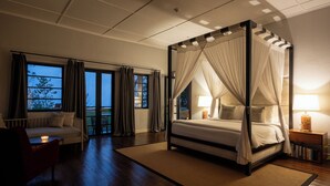 Ein Bett mit Baldachin, ein Nachttisch mit Lampe, eine Couch und ein Blick ins Freie durch ein großes Fenster.