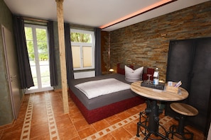 A bedroom with a bed, a round table with a chair, a brick wall, and a window with curtains.