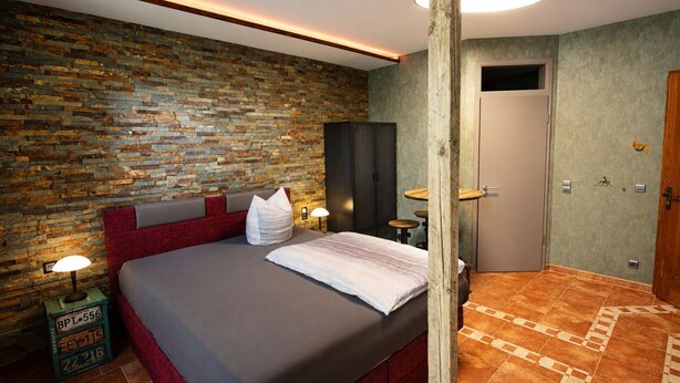 Un dormitorio con pared de piedra, una cama con cabecera roja, un pilar de madera y una puerta.