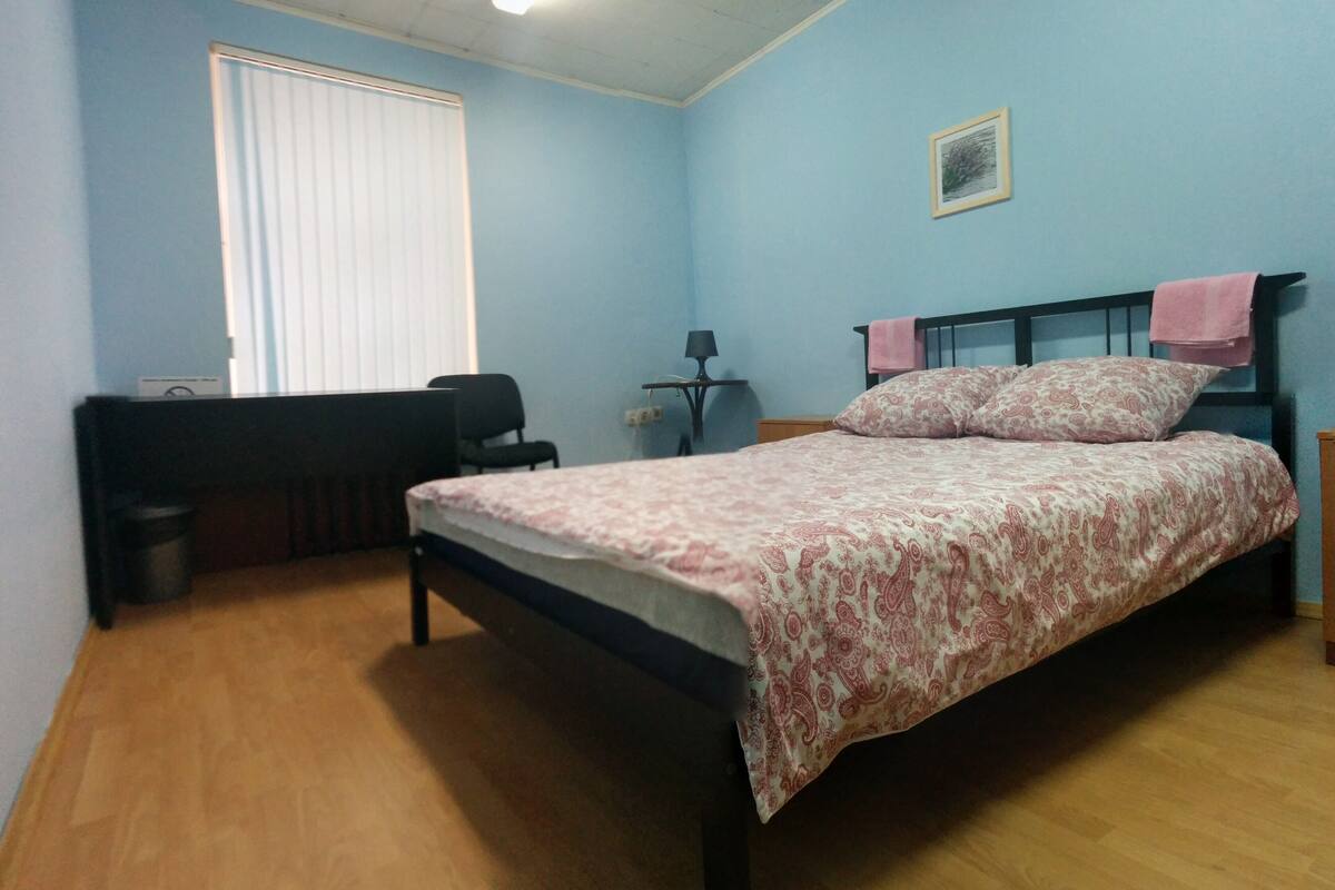 A bedroom with a bed, a desk, a chair, a lamp, and a window with blinds.
