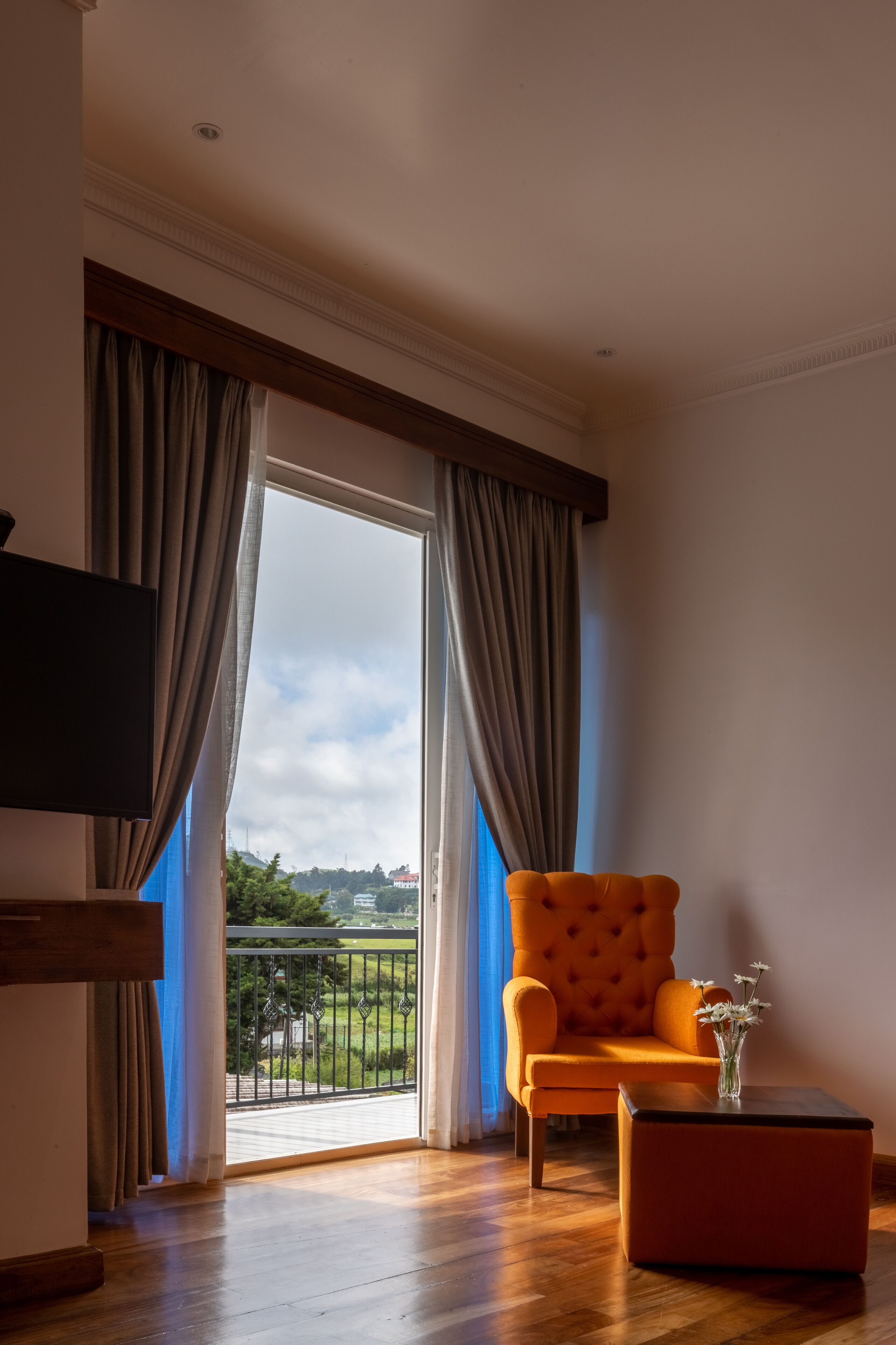A room with a wooden floor, a large window with curtains, a television, an orange armchair, and a small table with a vase.