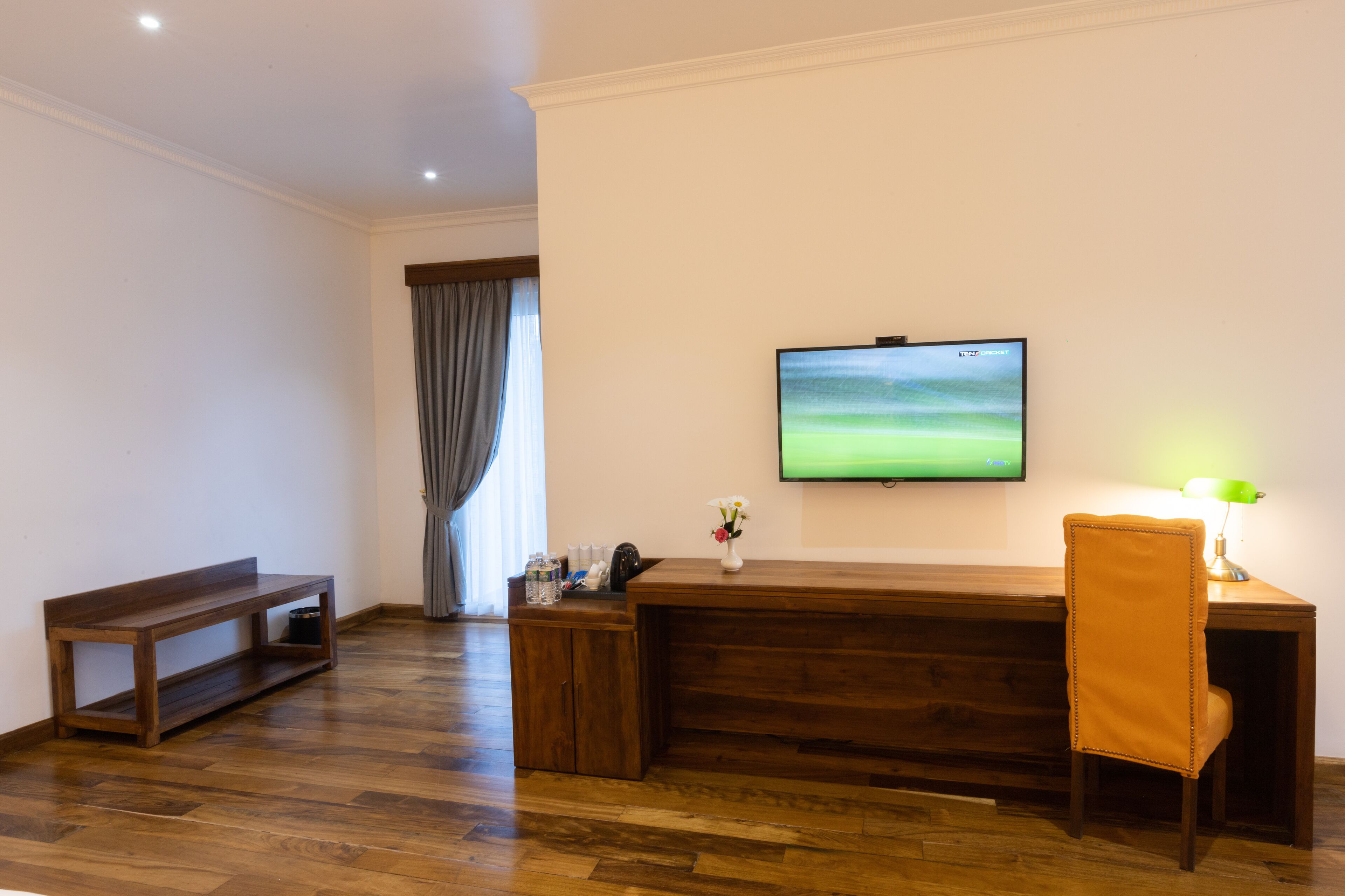 A room with a wooden desk, a chair, a television mounted on the wall, and a bench.