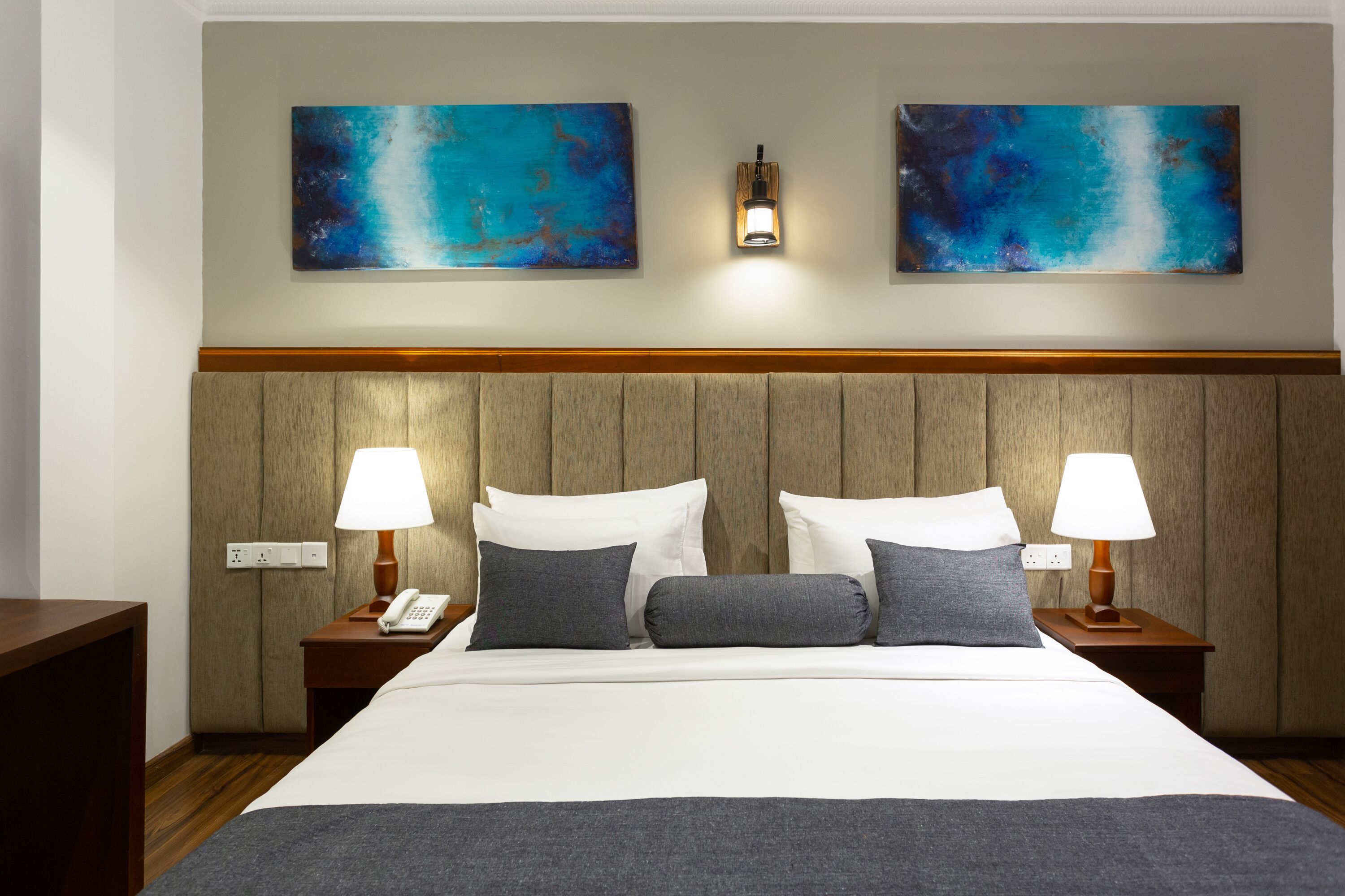 Habitación de hotel con una cama grande, dos mesitas de noche con lámparas, un cabecero de madera y dos cuadros abstractos en la pared.