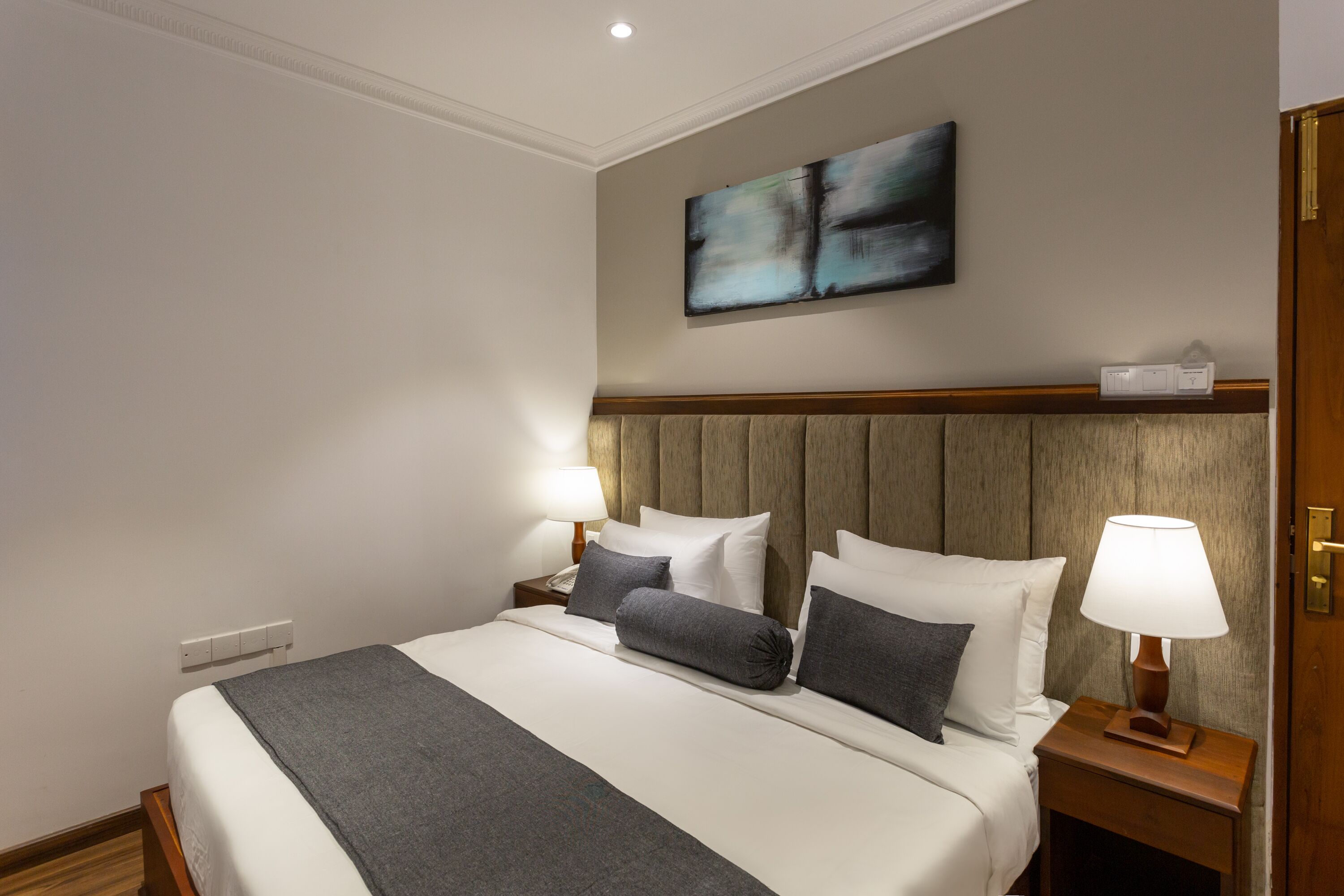 Une chambre d’hôtel avec un grand lit, deux tables de chevet, une tête de lit en bois, un tableau abstrait accroché au mur et une petite table de nuit avec une lampe.
