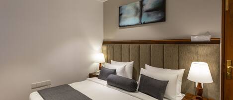 Une chambre d’hôtel avec un grand lit, deux tables de chevet, une tête de lit en bois, un tableau abstrait accroché au mur et une petite table de nuit avec une lampe.
