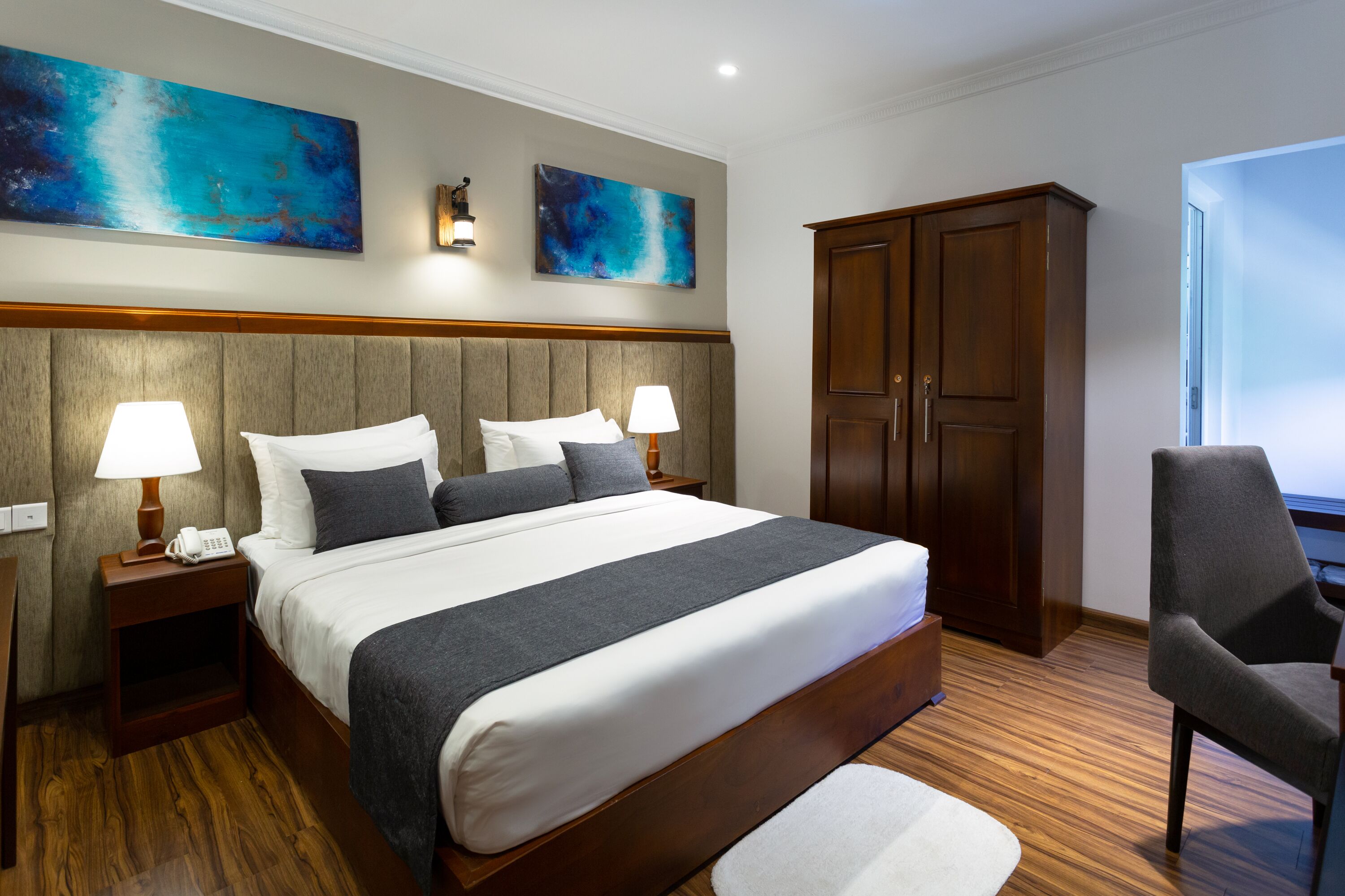 Habitación de hotel con una cama grande, cabecera de madera, dos lámparas de noche, una mesita de noche, un armario y una silla.