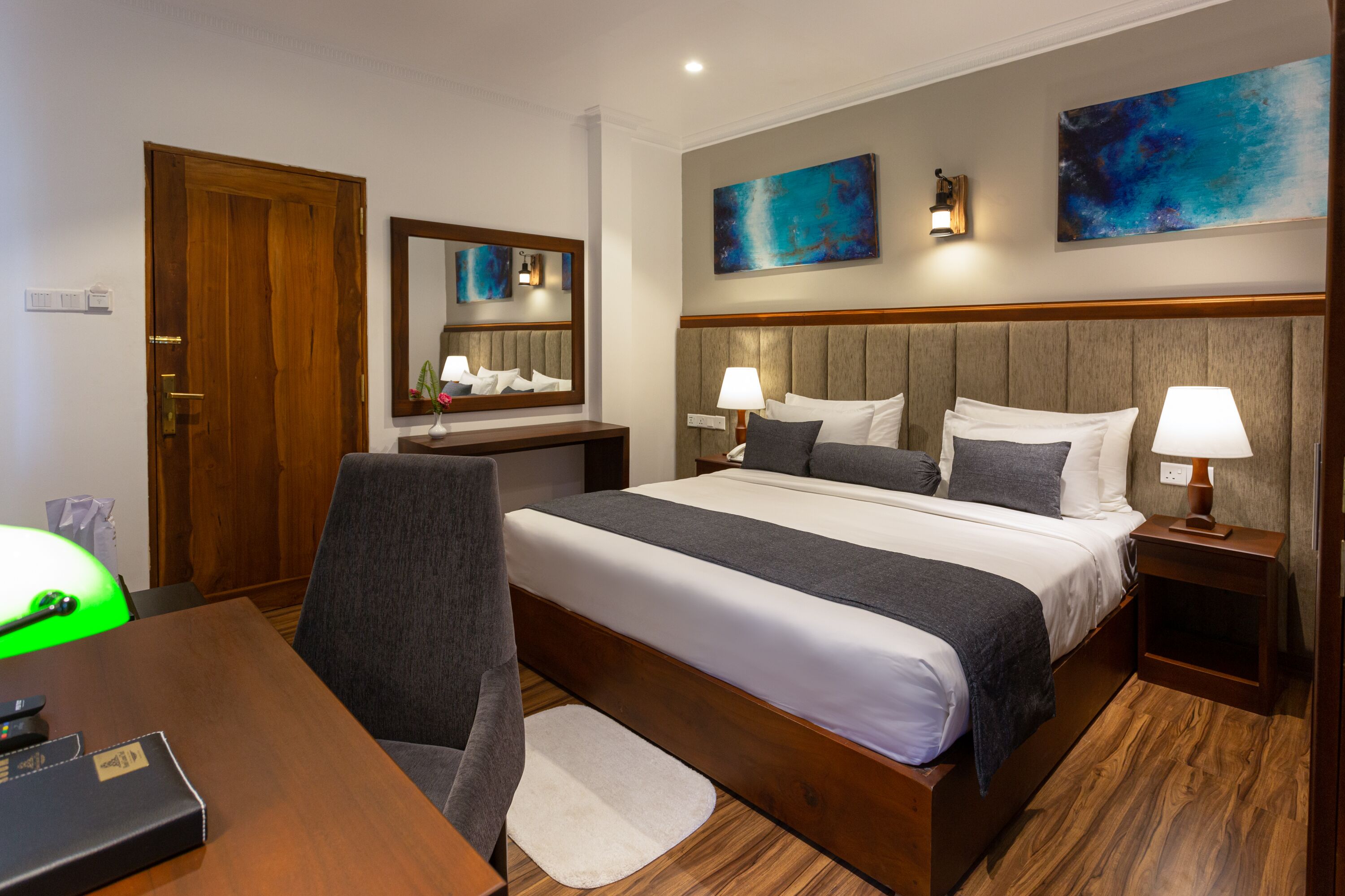 A hotel room with a large bed, two bedside tables with lamps, a desk with a chair, a mirror, and artwork on the walls.