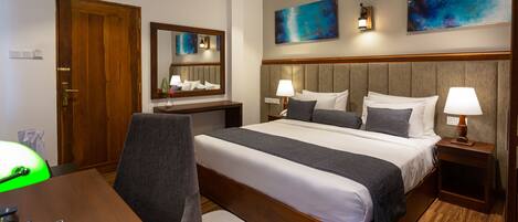 A hotel room with a large bed, two bedside tables with lamps, a desk with a chair, a mirror, and artwork on the walls.
